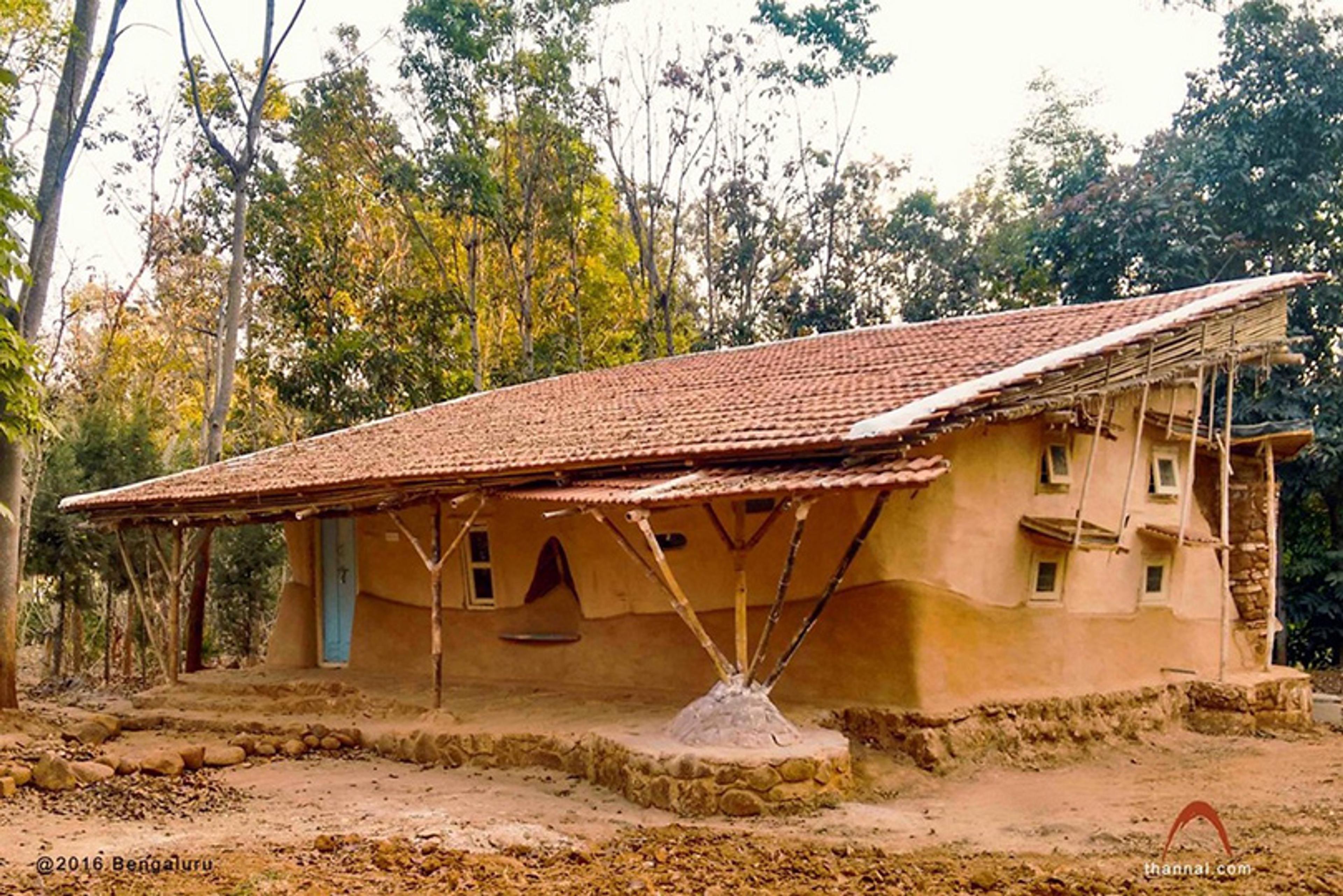 Photo of an earthen house with a sloped tiled roof supported by bamboo, set amidst trees and surrounded by rustic landscaping.