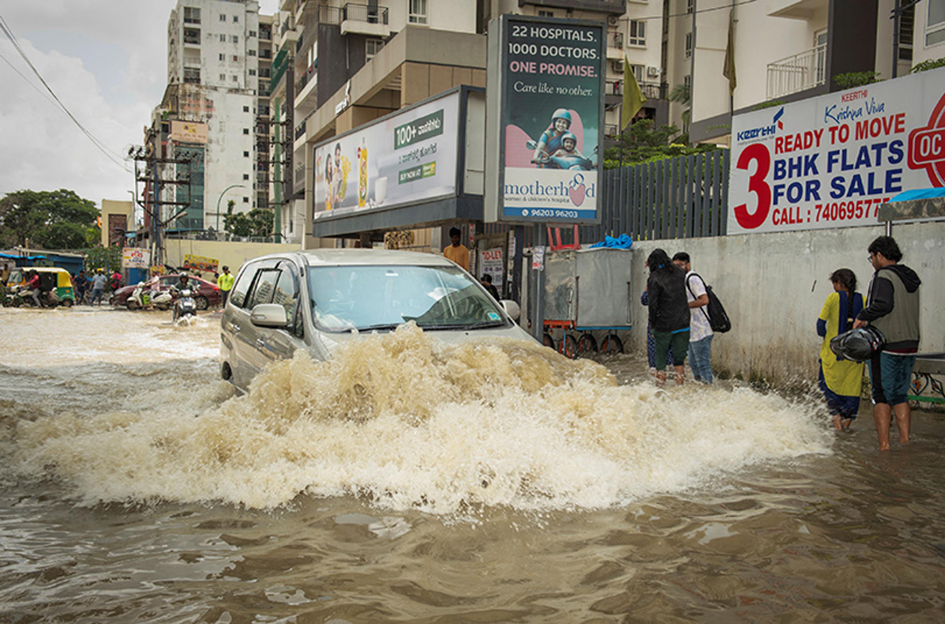 Photo of a car driving through a flooded street with people walking on the side in a city with advertising billboards.