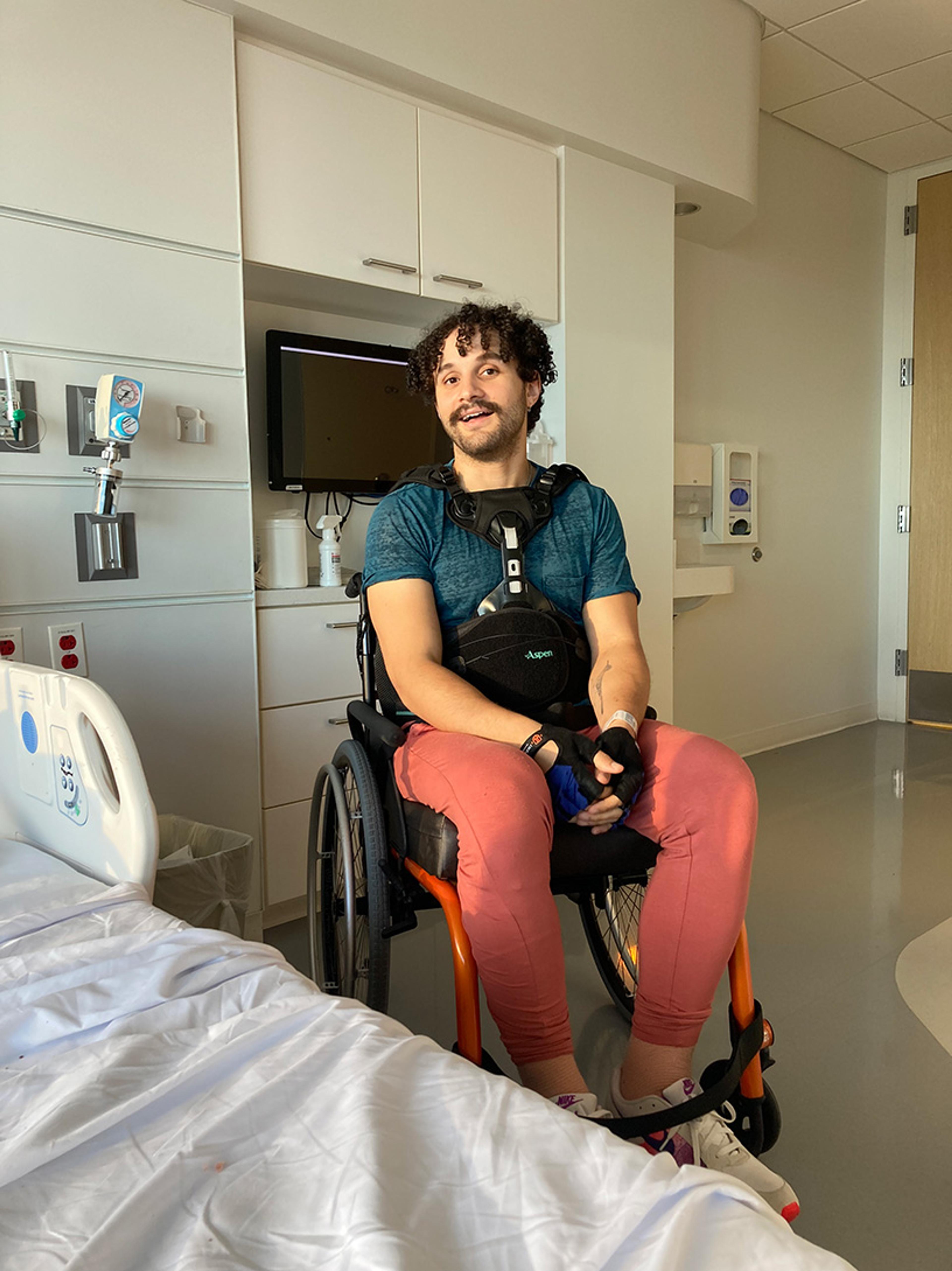 A man in a wheelchair smiling in a hospital room with a bed and medical equipment.