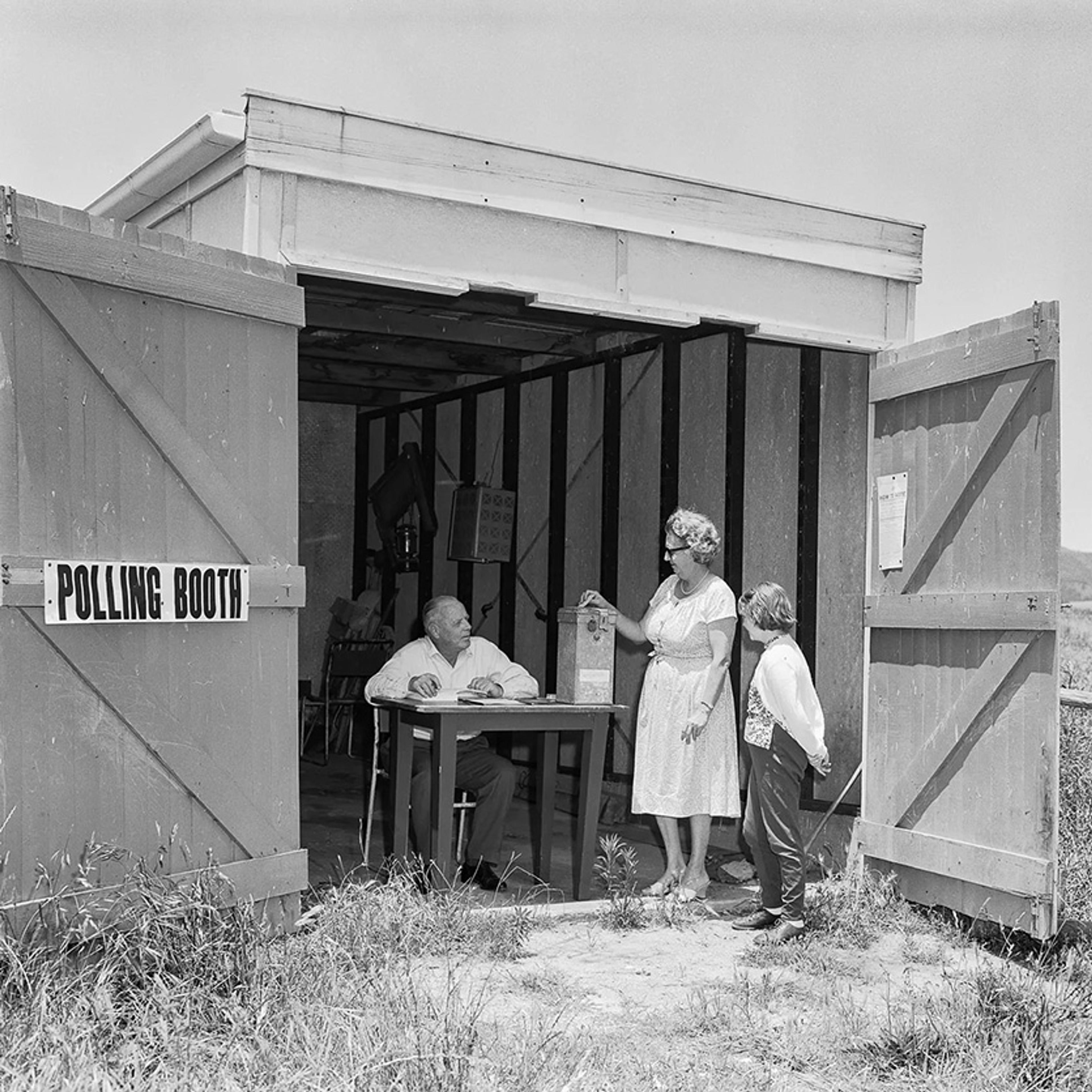 Black and white photo of a polling booth in a shed where a woman votes as a man and child watch.