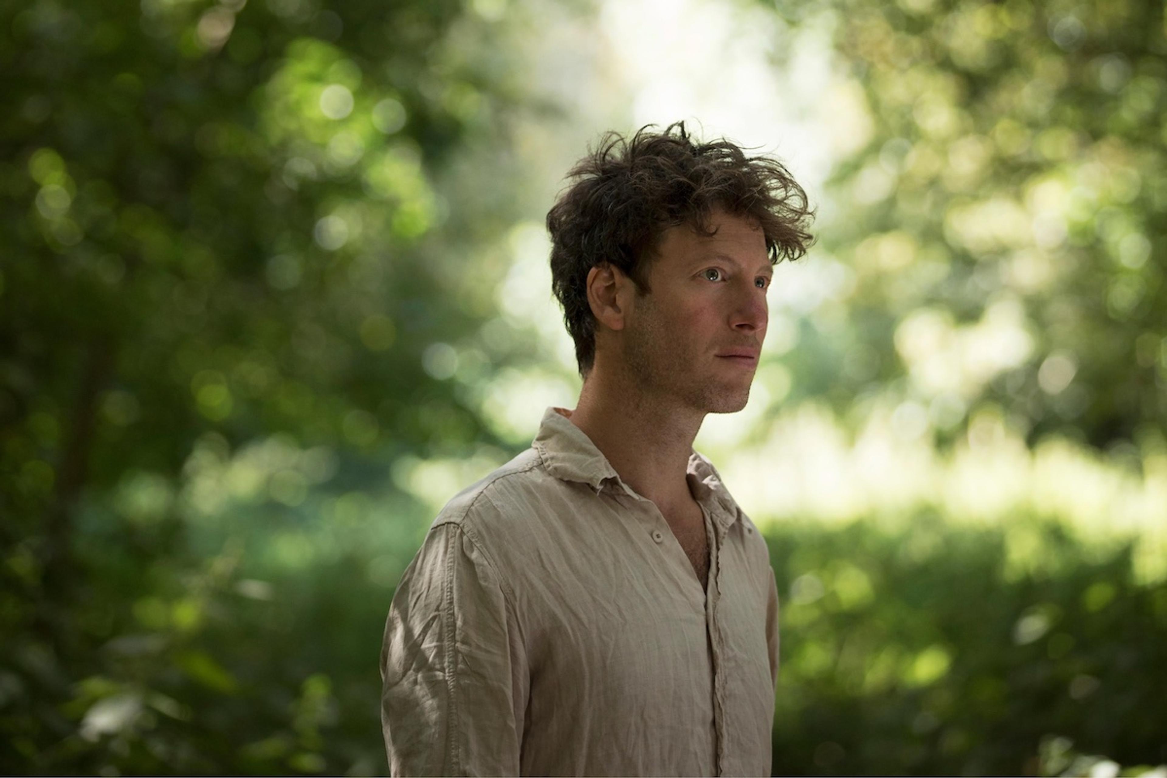 Photo of a man standing outdoors, surrounded by blurred green foliage.