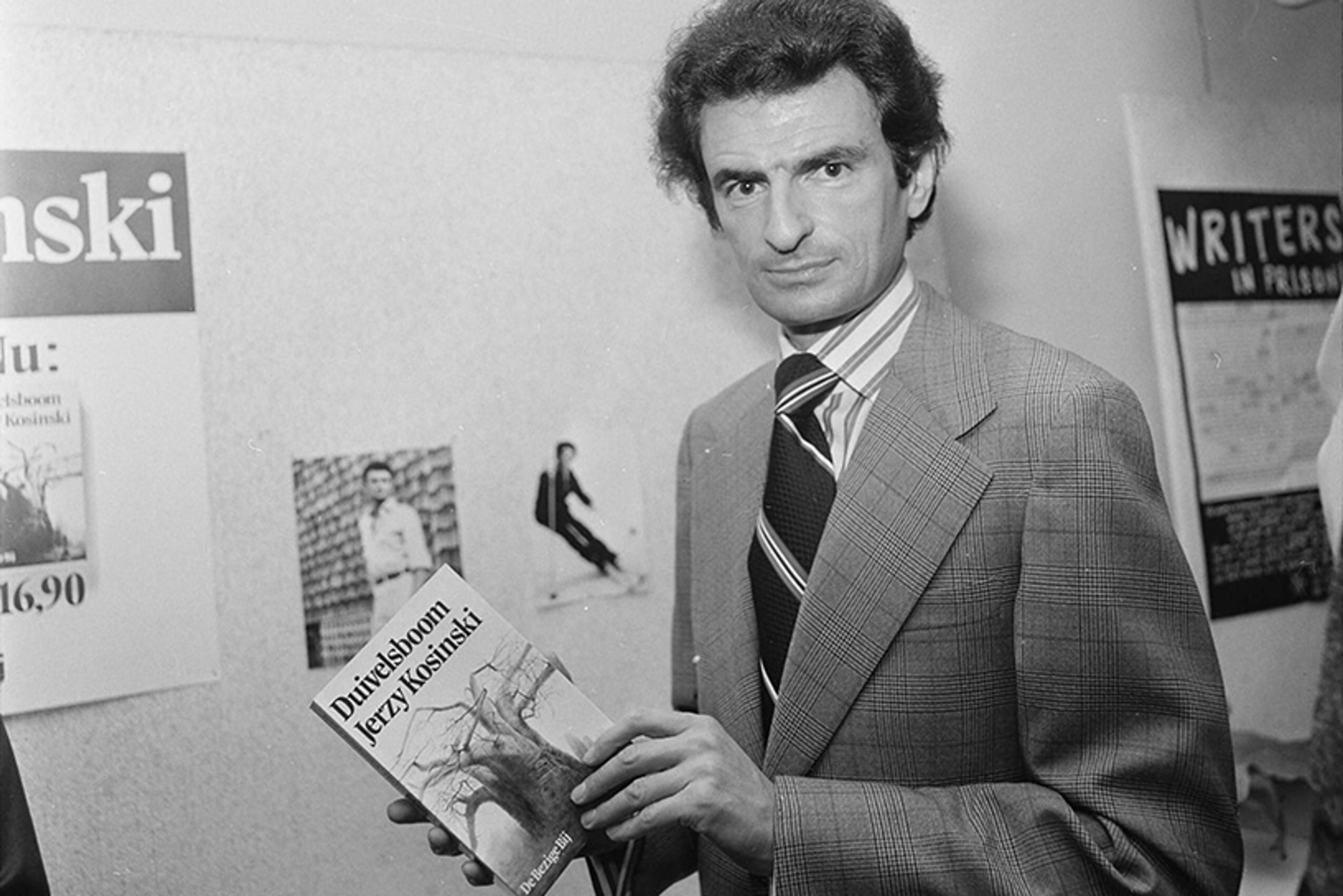Black and white photo of a man in a suit holding a book in a room with posters on the wall.