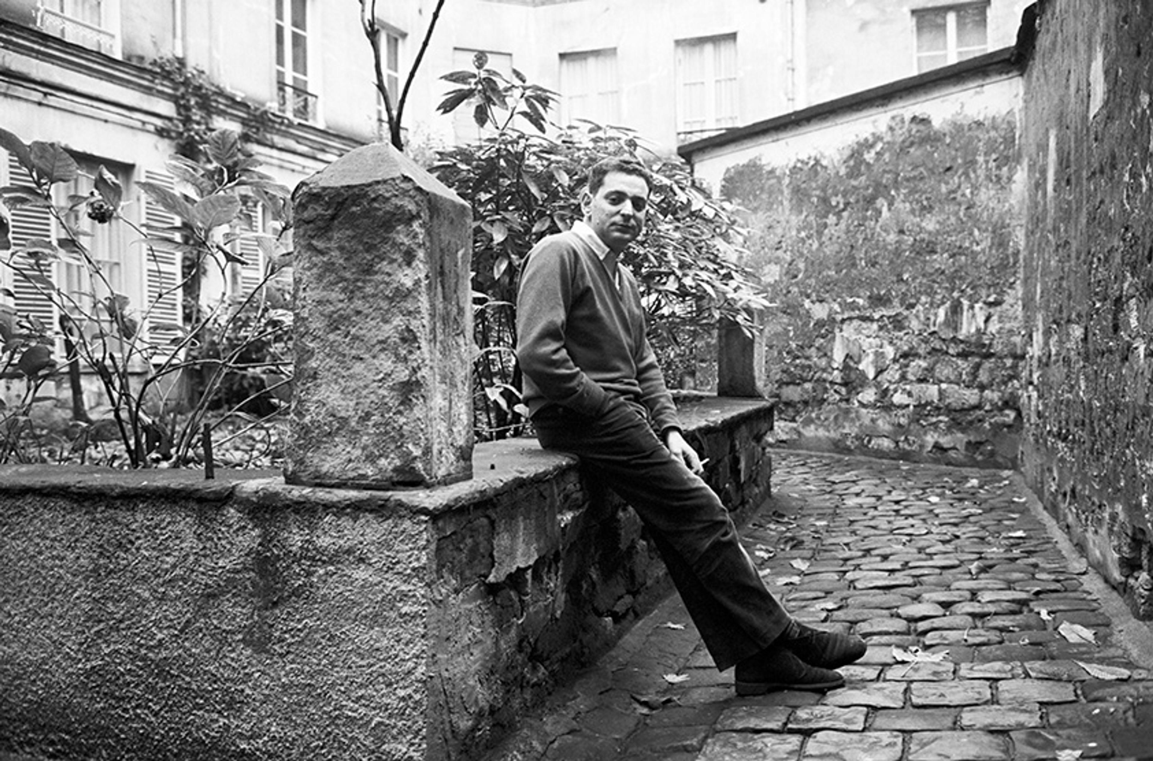 Black and white photo of a man sitting on a stone ledge in a cobblestone courtyard with surrounding buildings and plants.