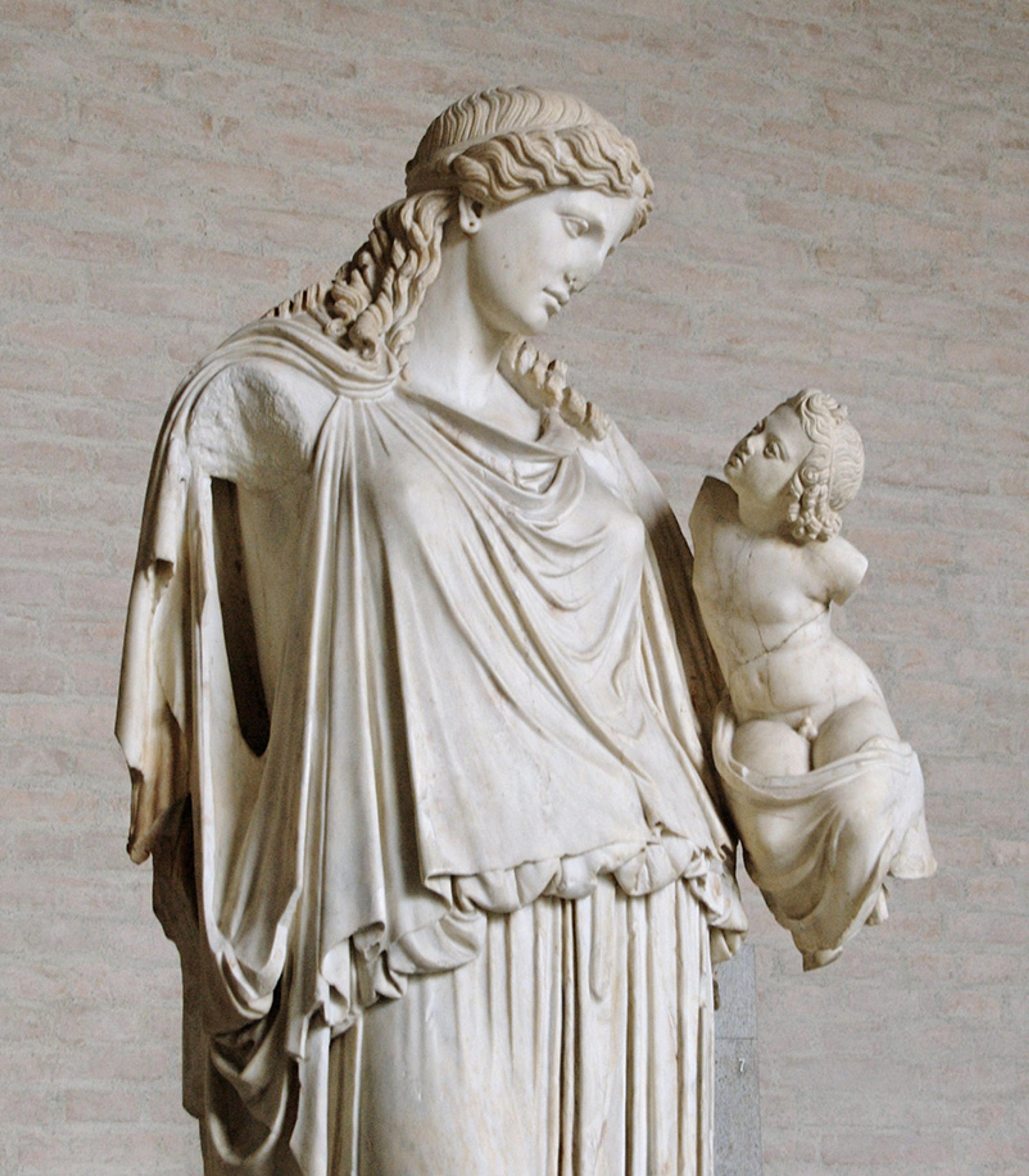 Ancient marble statue depicting a robed woman and a wingless cherub against a stone wall background.