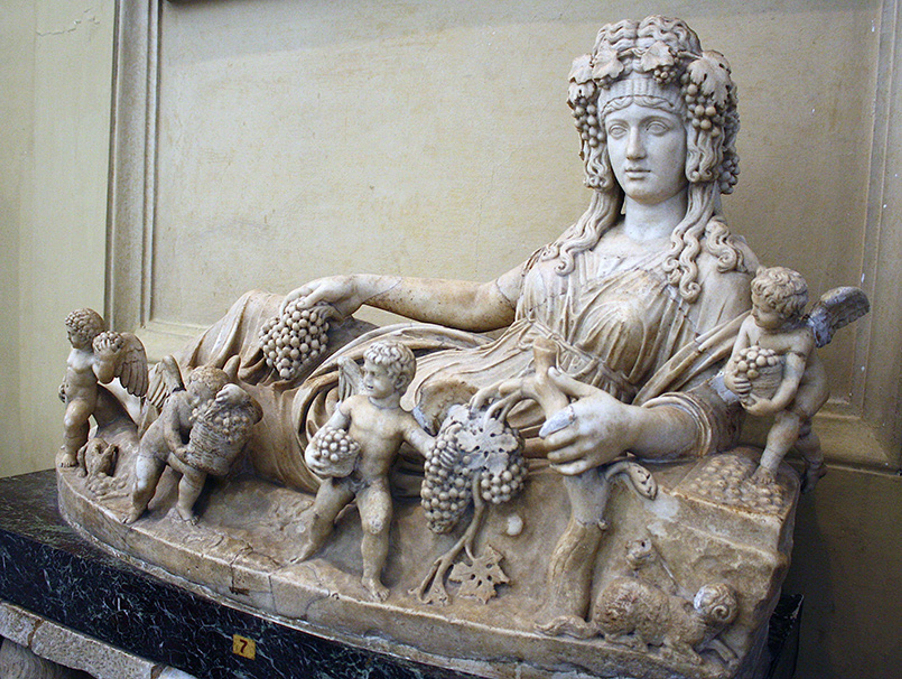 Marble sculpture of a reclining figure with grapevines and cherubs in a classical style, displayed on a dark marble base.
