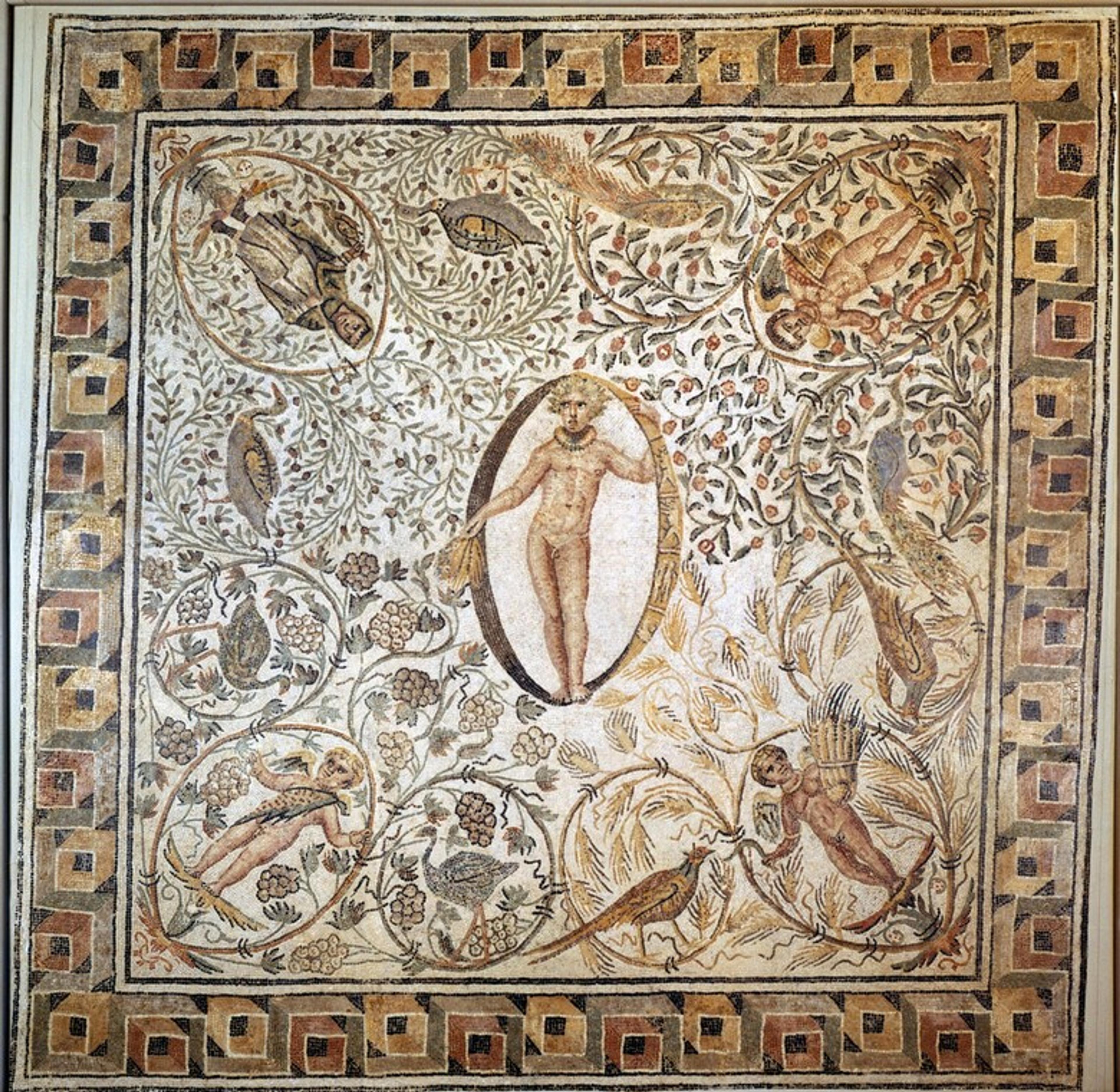 Ancient mosaic with a central nude figure surrounded by floral motifs and figures, bordered by geometric patterns.