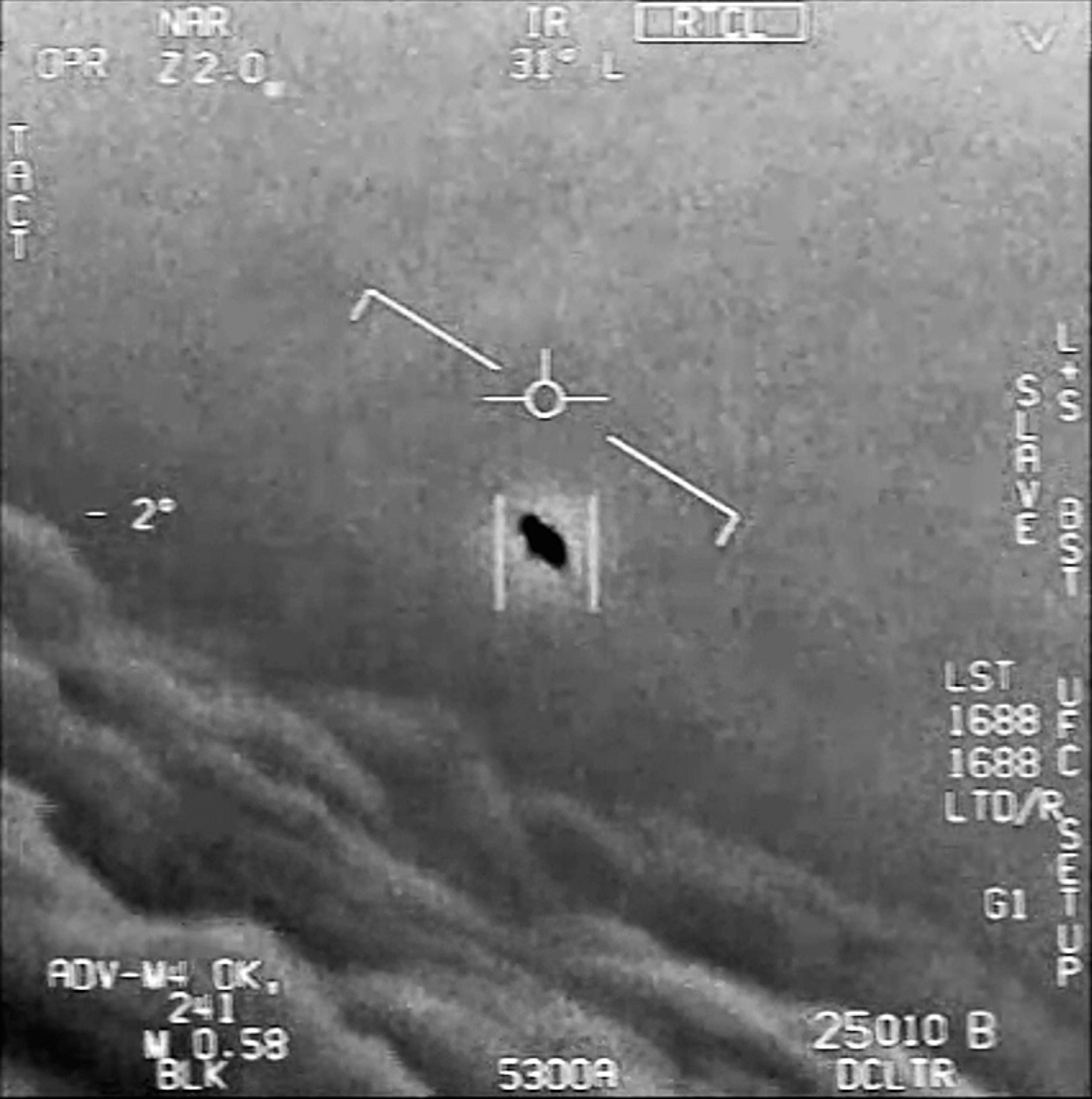 Black-and-white video still from an aircraft display showing an oval black object above clouds.