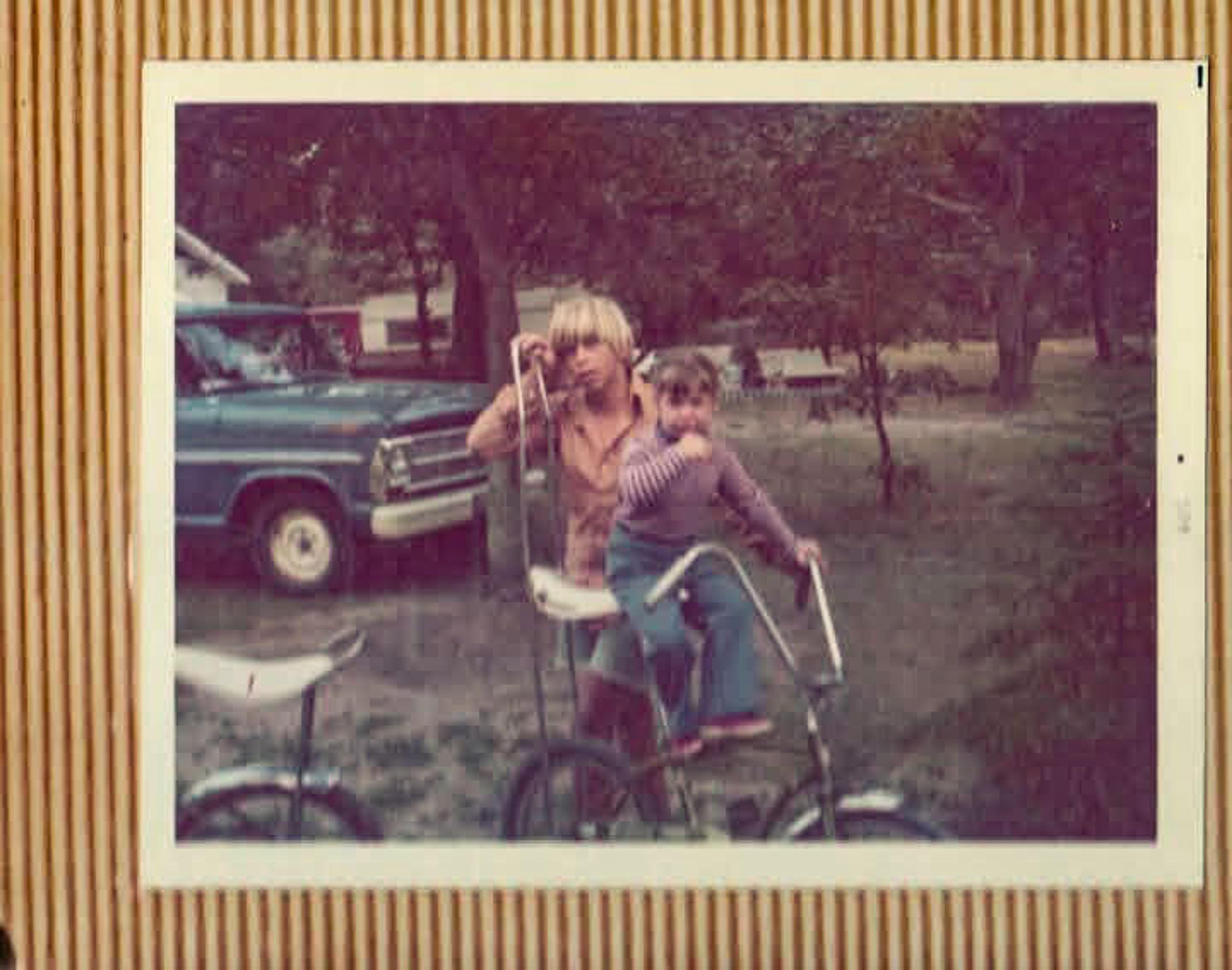 Vintage photo of two children with a bicycle, one seated, another standing, with an old truck and trees in the background.
