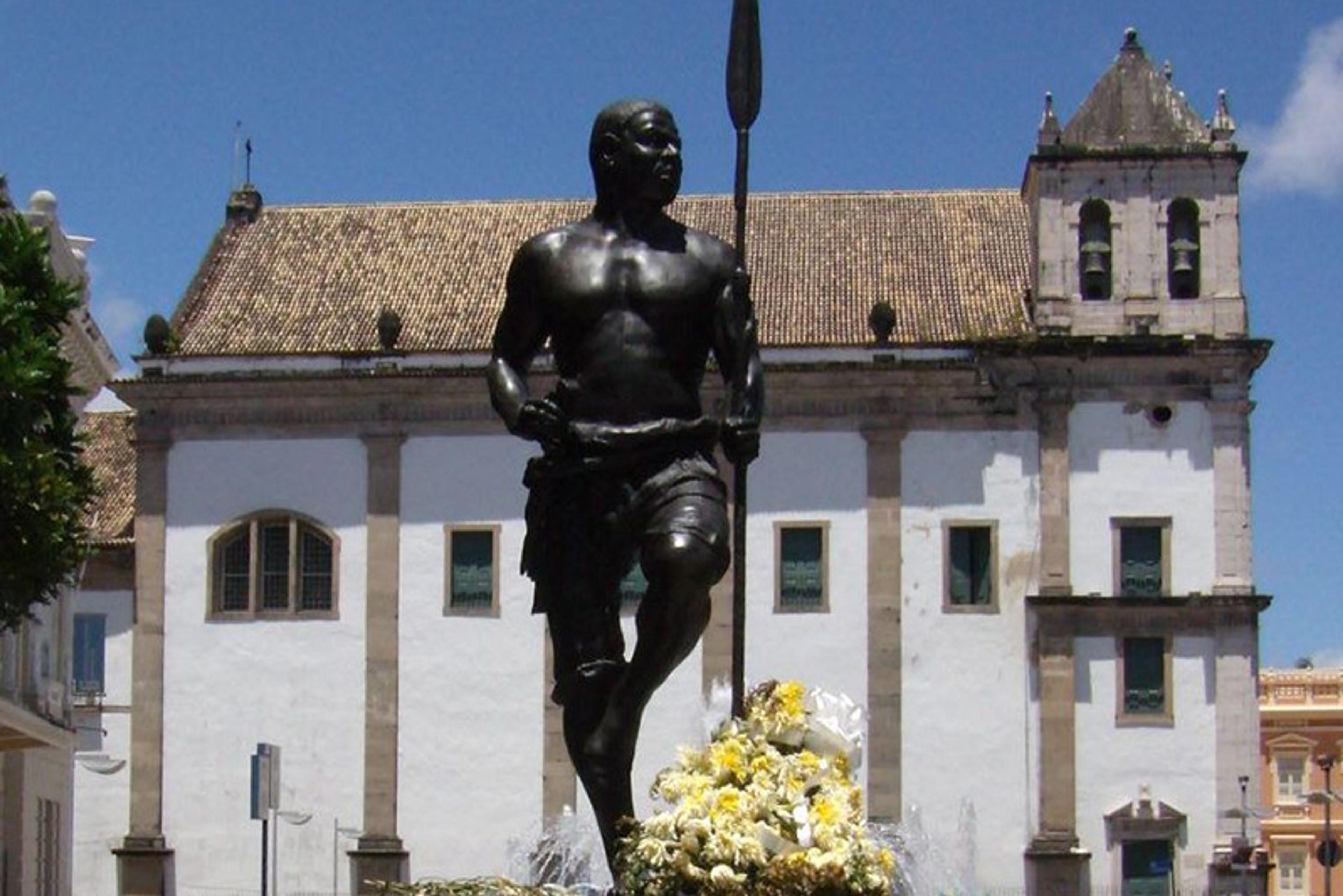 Photo of a statue of a man holding a spear in front of a historic building, with flowers at the base and a clear sky.