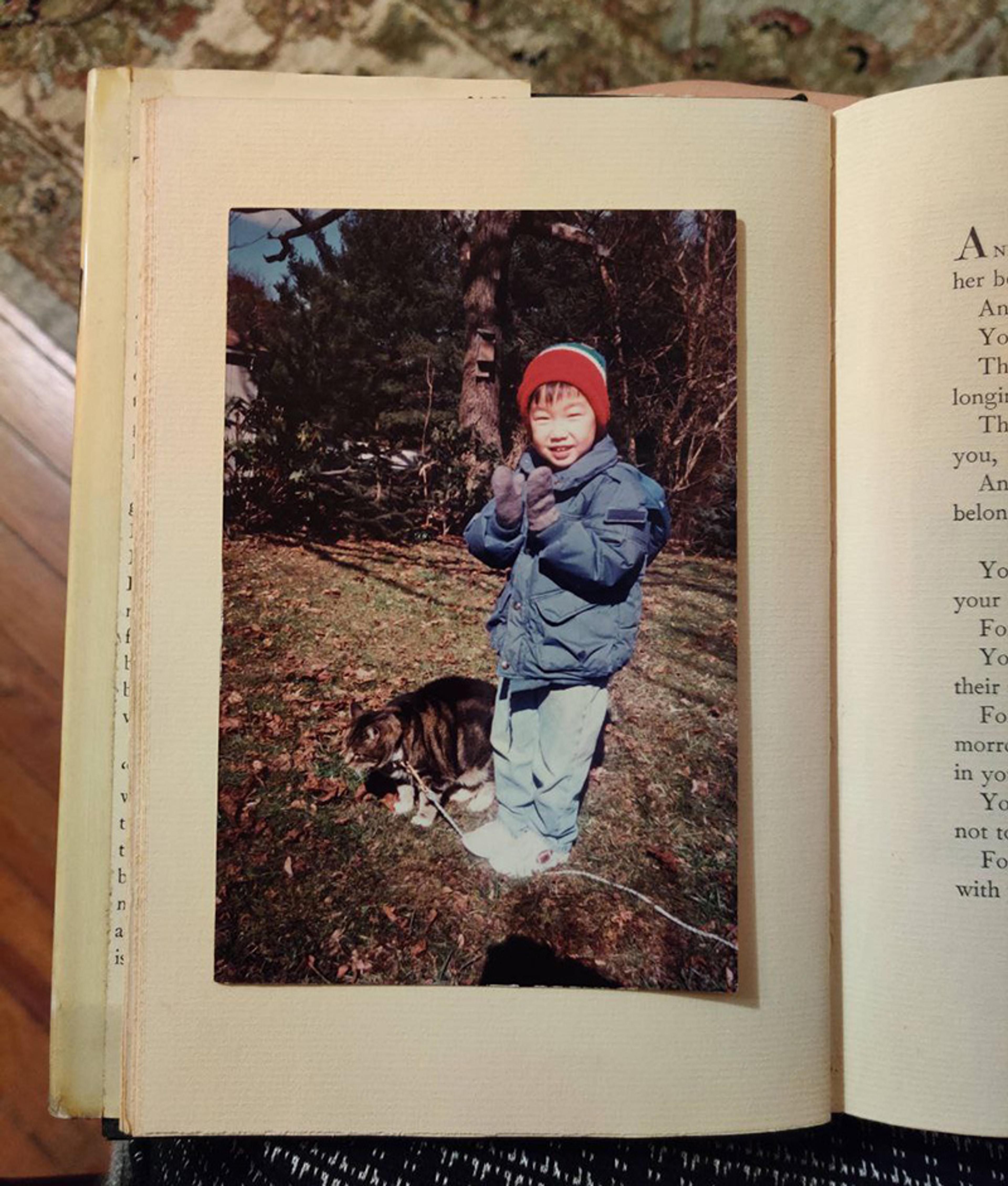 Photo of a child in winter clothes in a garden with a cat. The photo is placed inside an open book on a patterned surface.