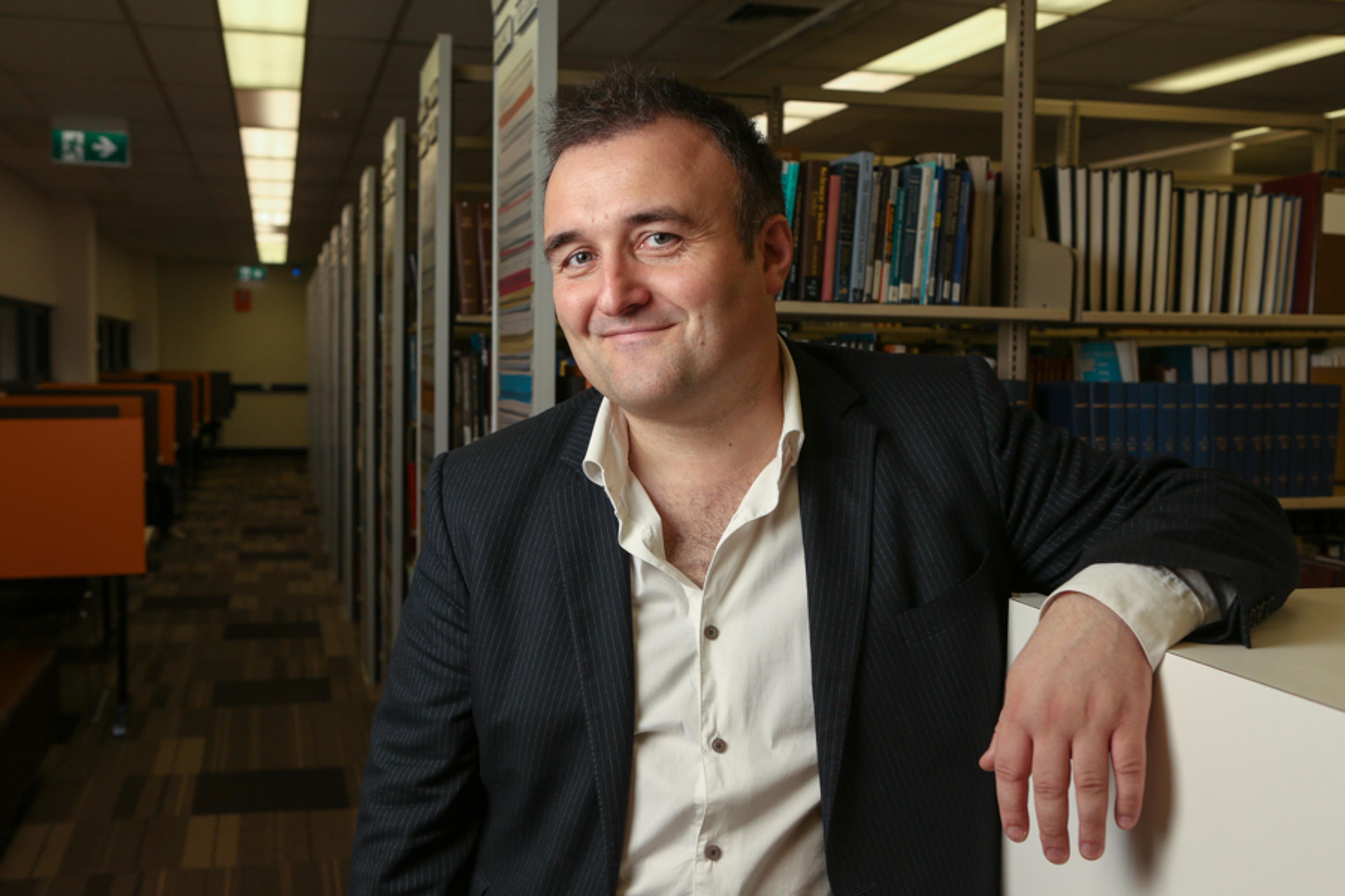 Photo of a man in a suit leaning on a library shelf lined with books, smiling at the camera in a well-lit library.