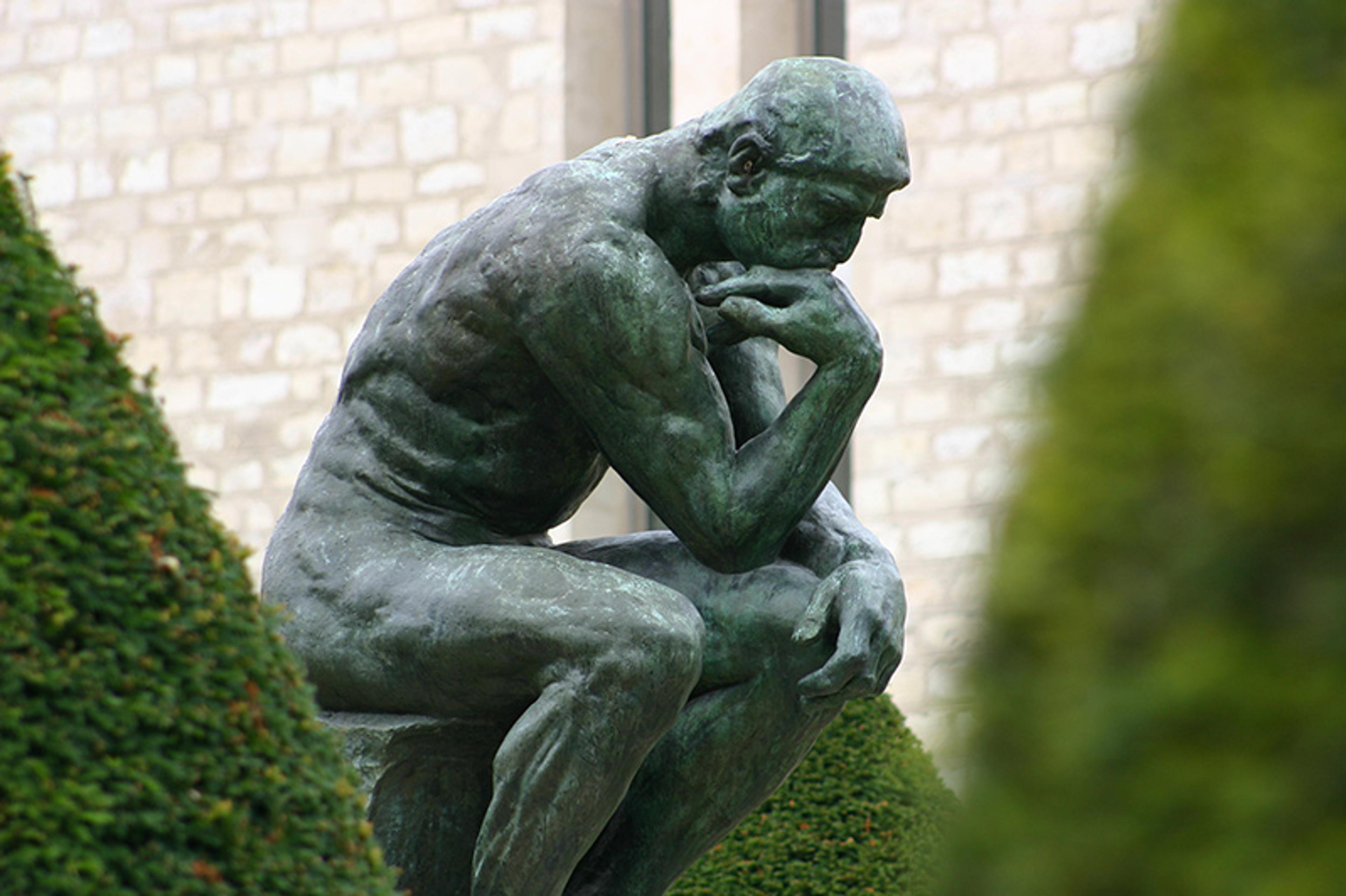 A bronze statue of a nude man seated, leaning forward, his right elbow placed on his left thigh, with the back of his right hand supporting his chin in a posture of contemplation, surrounded by green hedges and a stone building backdrop