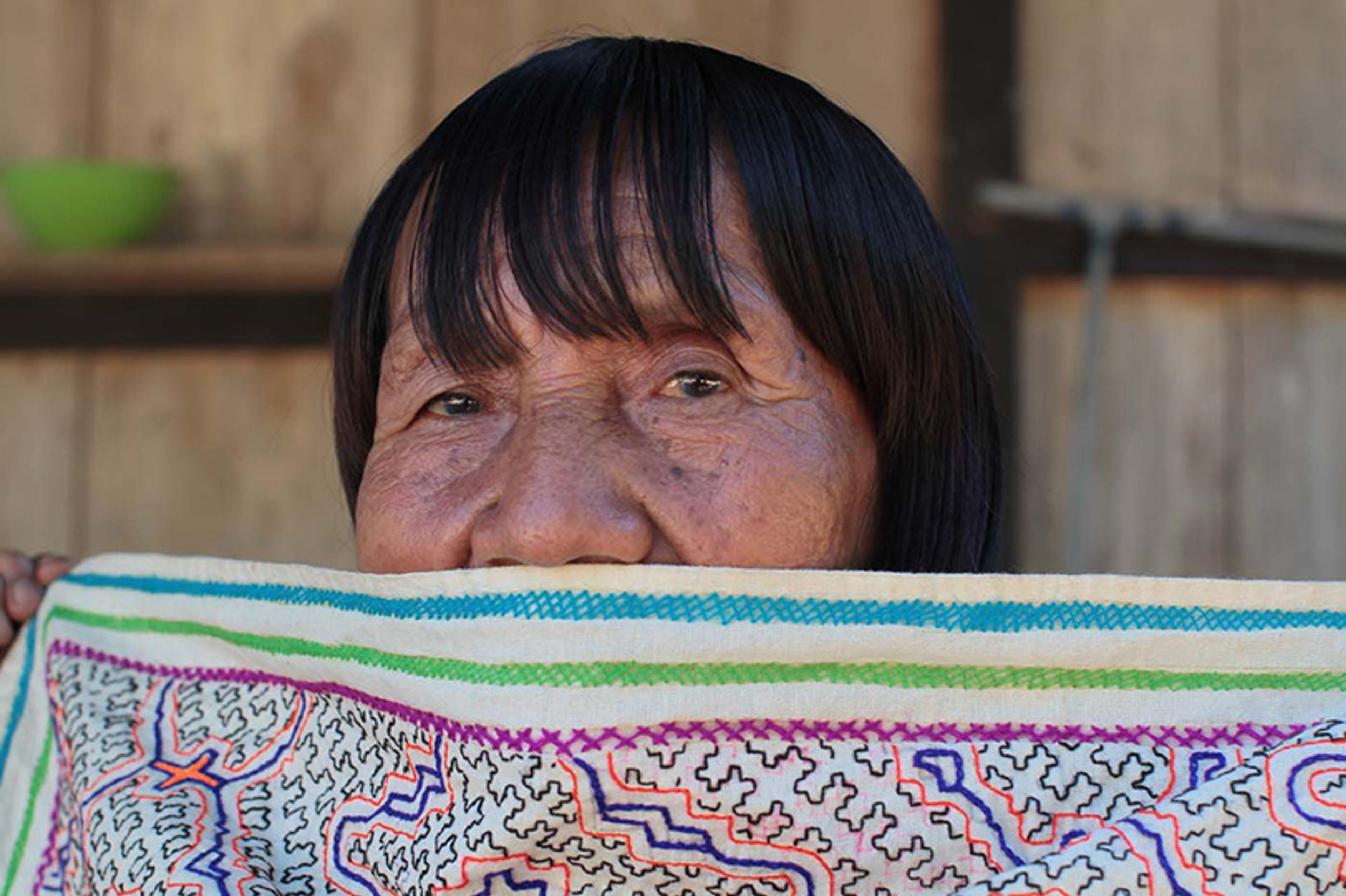 Photo of a woman’s face with the lower half of her face obscured by a colourful embroidered fabric, set against a wooden background.