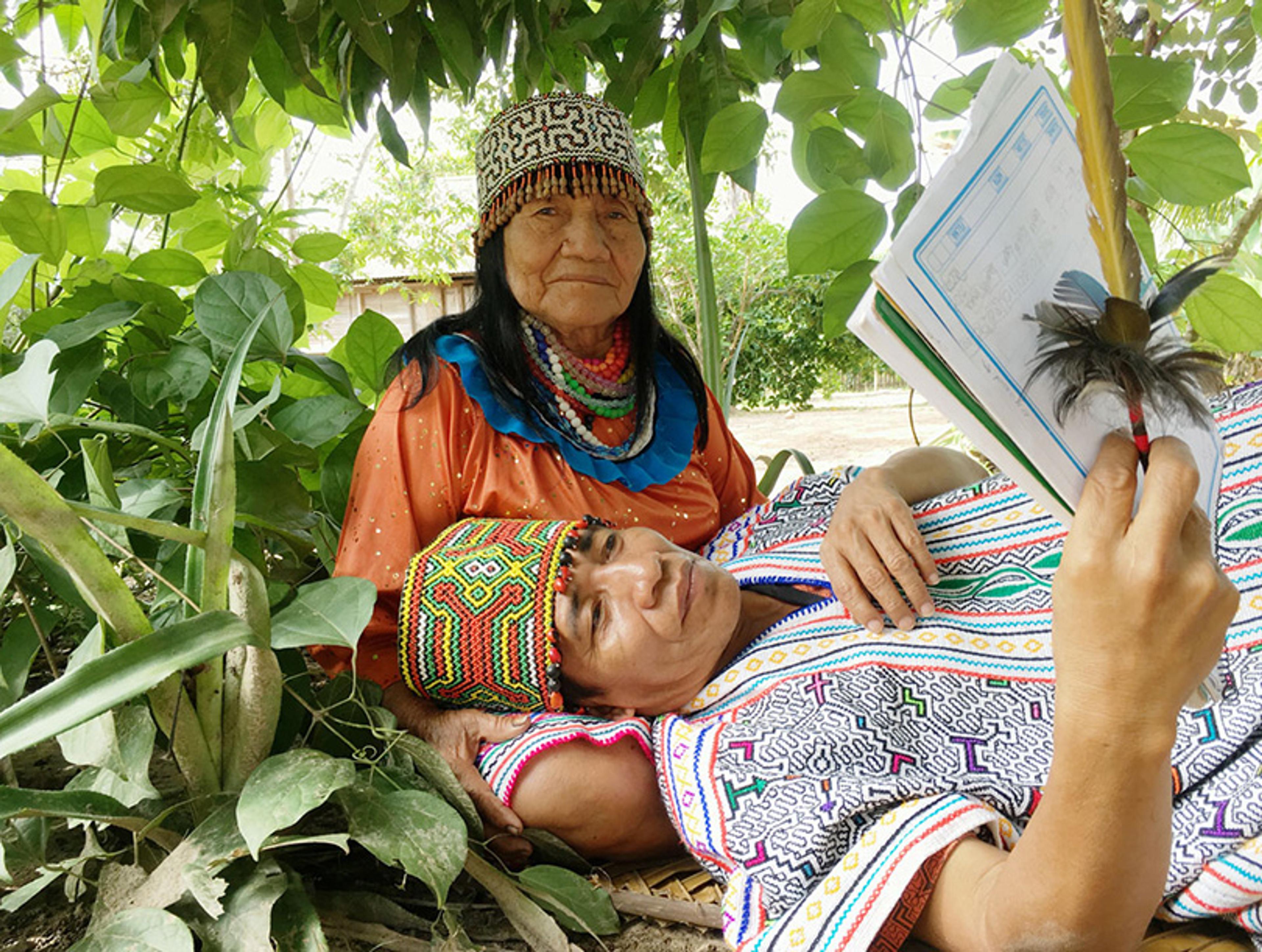 Photo of a woman sitting on the ground with a man lying on her lap while reading and holding a feather. They wear traditional clothing and are surrounded by green leaves