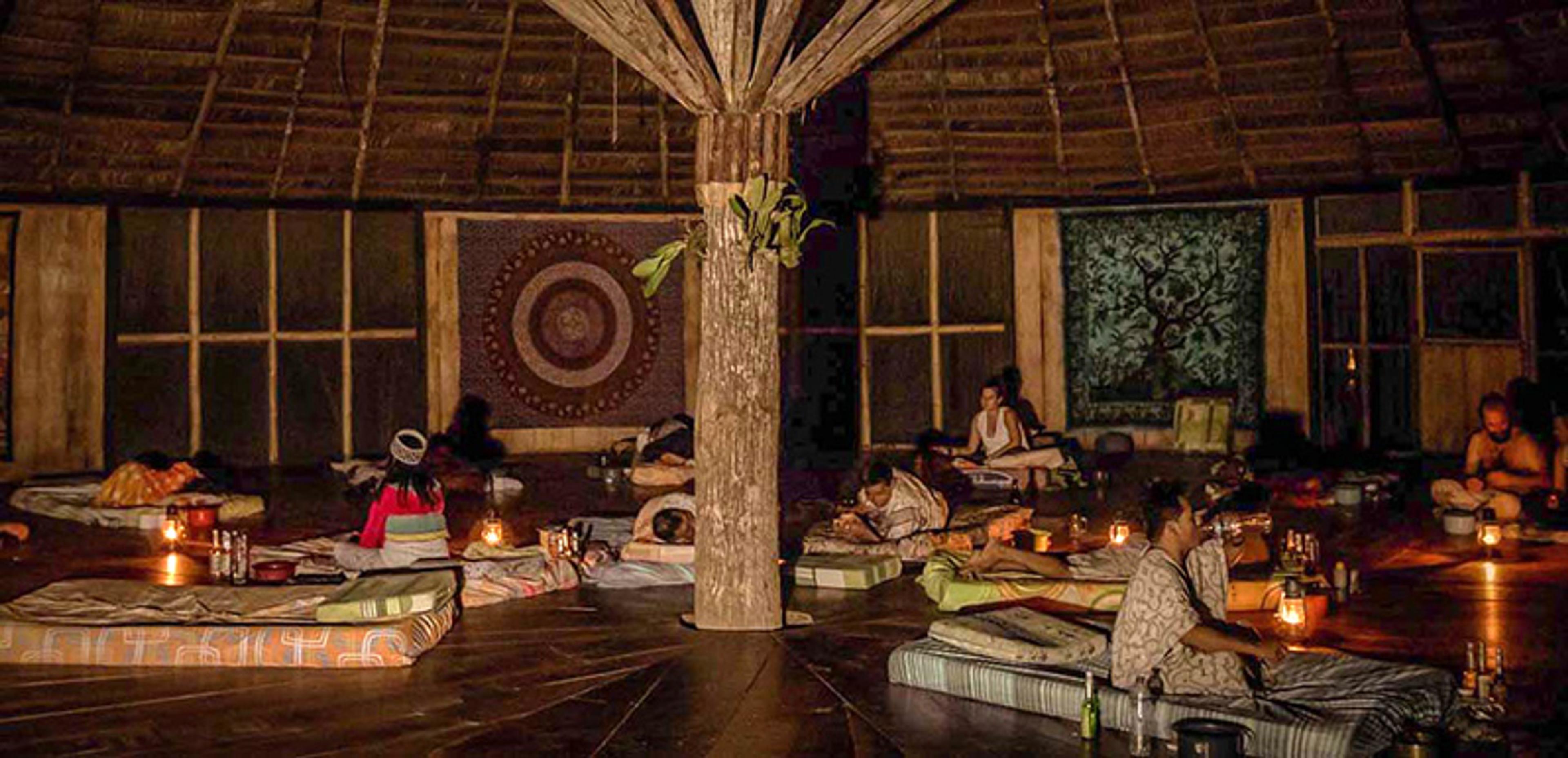 Photo of a dimly lit room with people sitting on mattresses around a central wooden pole, tapestries on the walls.