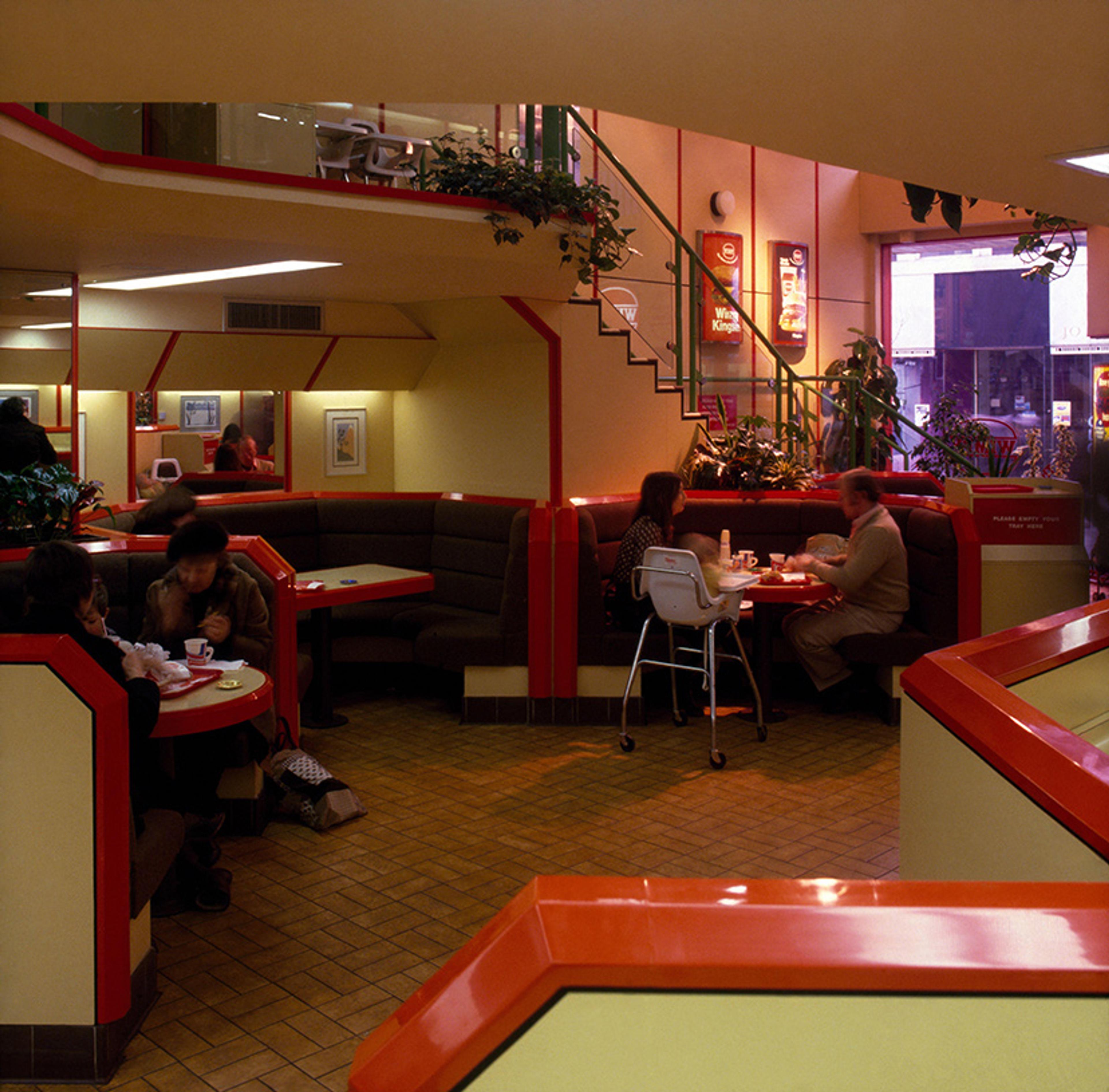 A retro fast-food restaurant interior with red booths and chairs, a staircase, and people dining.