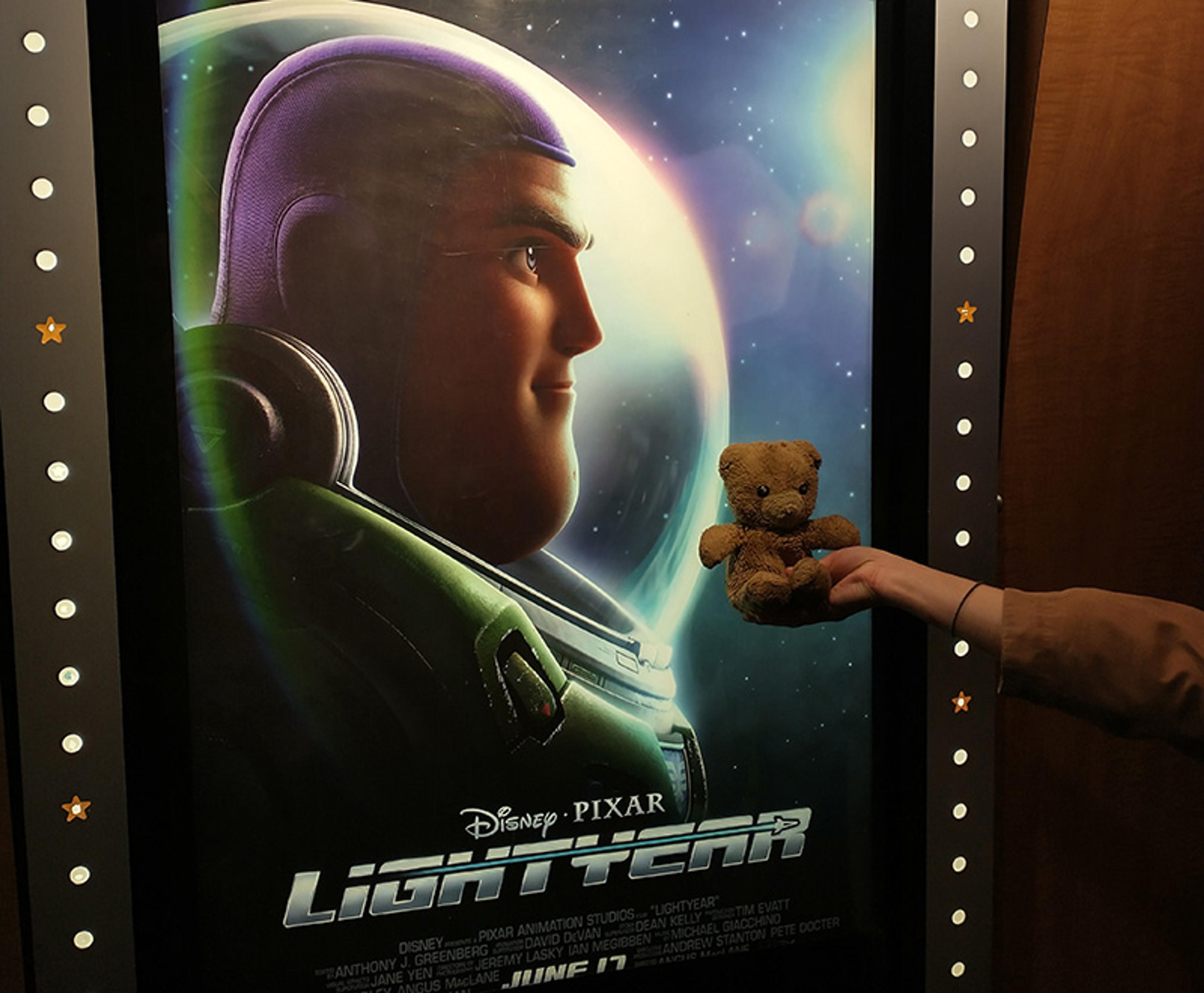 A “Lightyear” movie poster with Buzz Lightyear. A person holds a small teddy bear next to the poster.