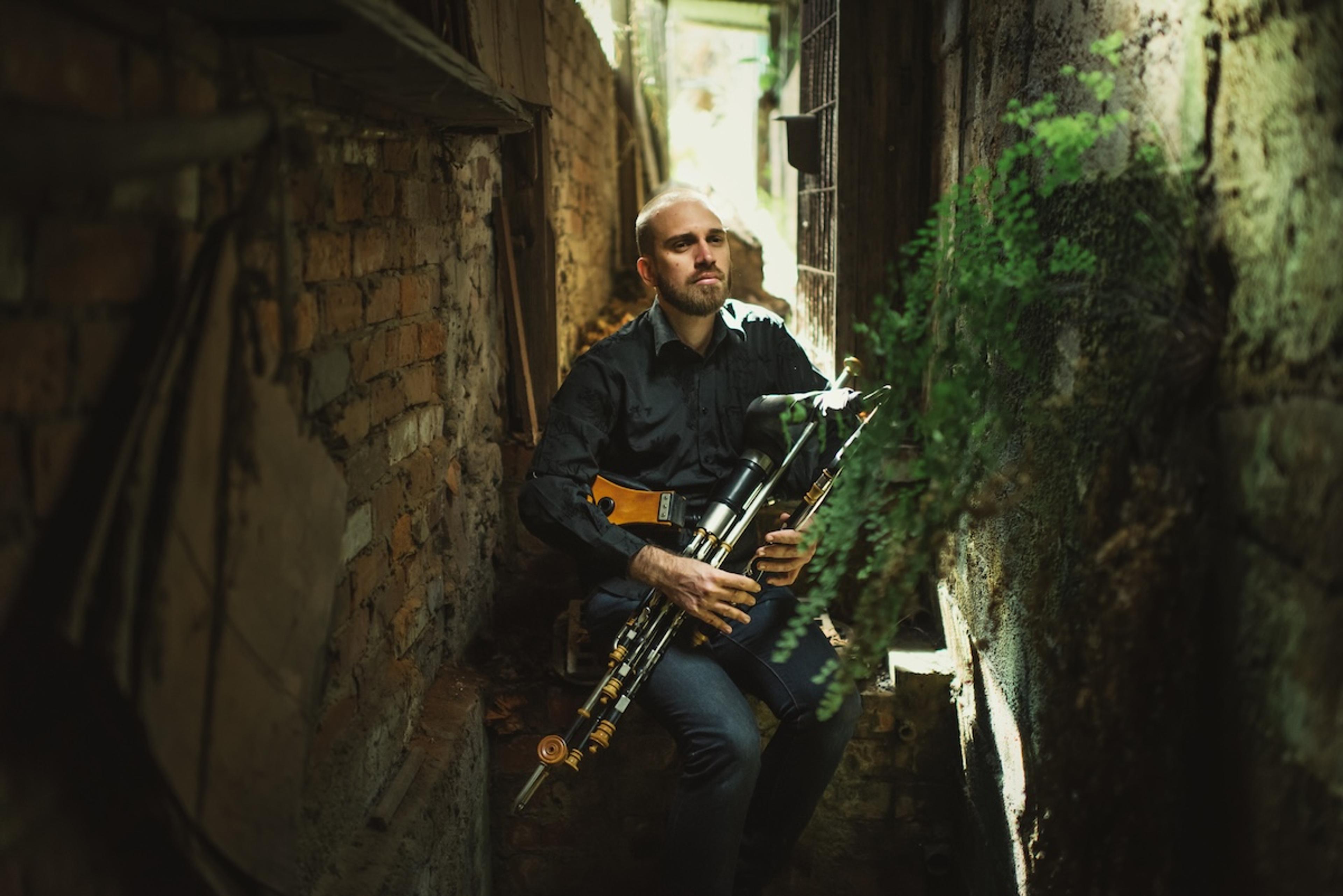 Photo of a man playing uilleann pipes in a narrow, dimly lit alley with brick walls and some greenery.