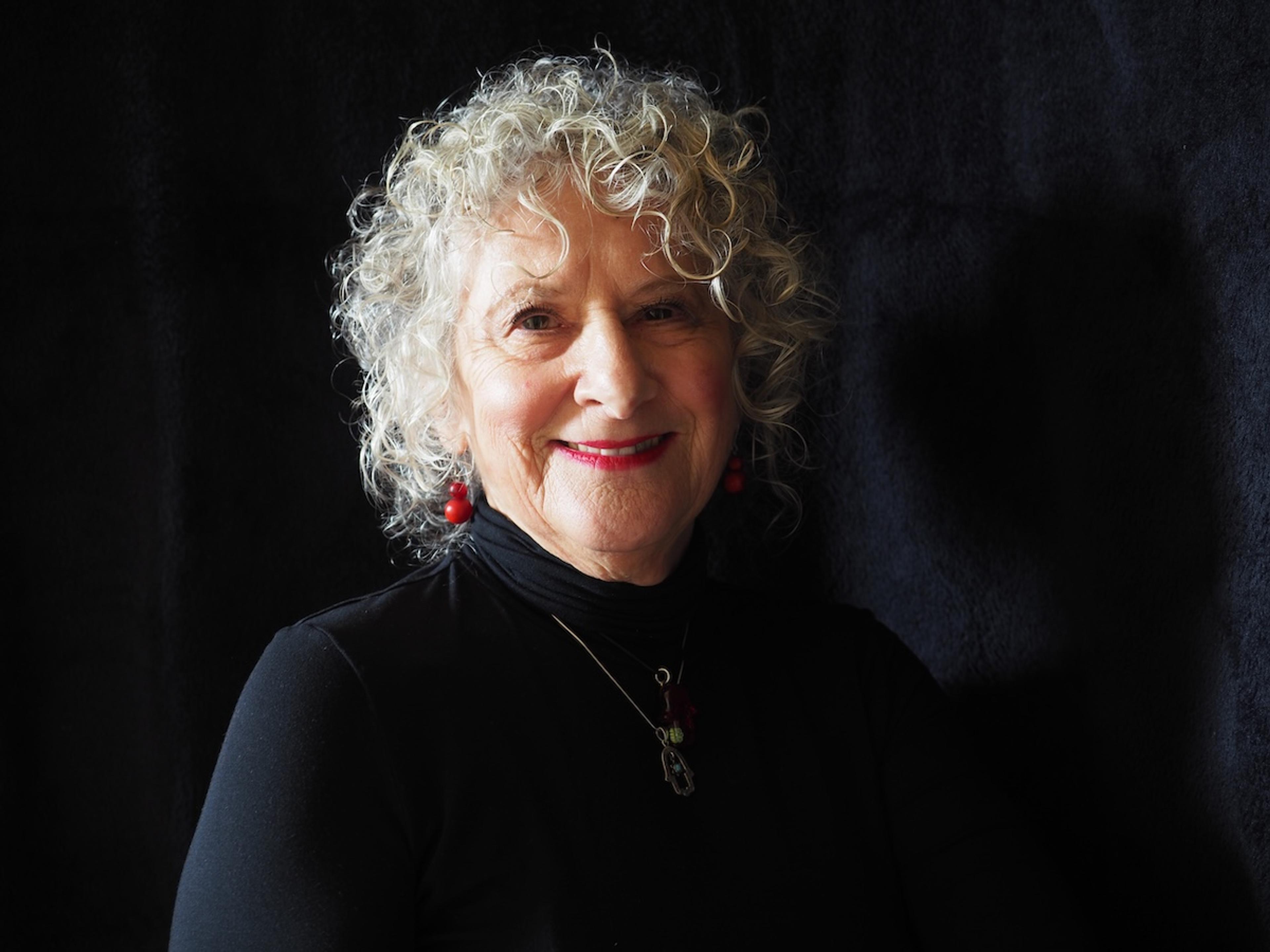 Photo of a smiling woman with curly white hair wearing a black top and red earrings against a dark background.