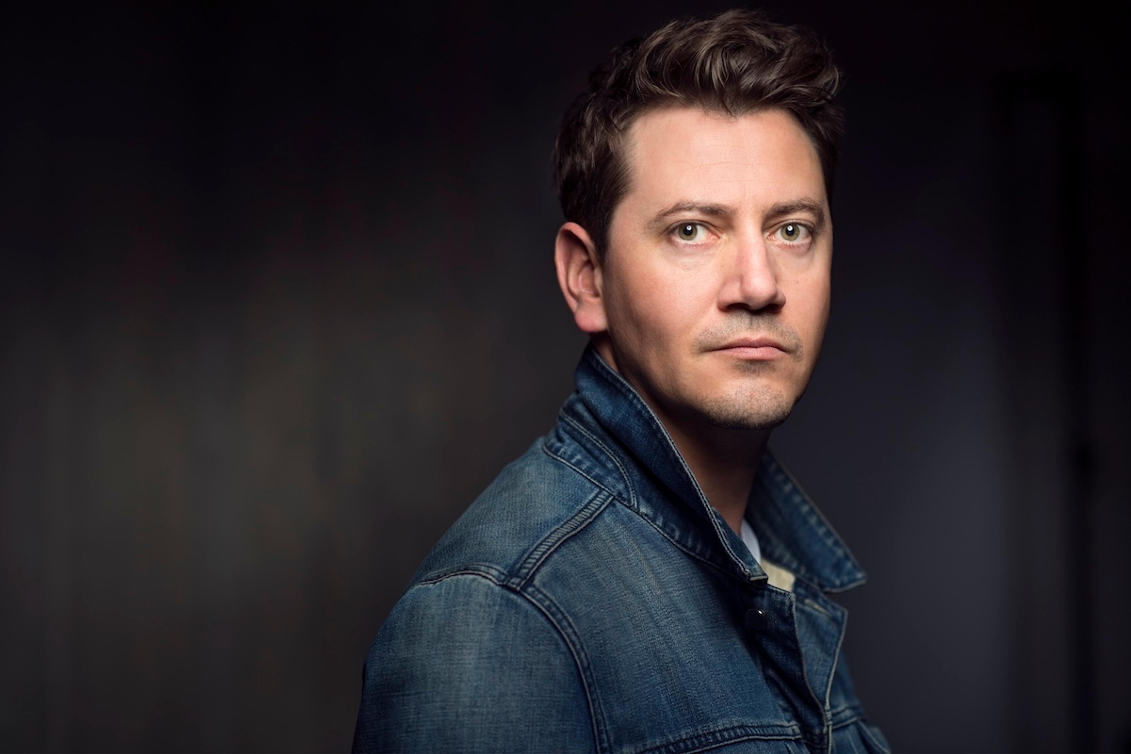 Photo of a man in a denim jacket with a neutral expression against a dark background.