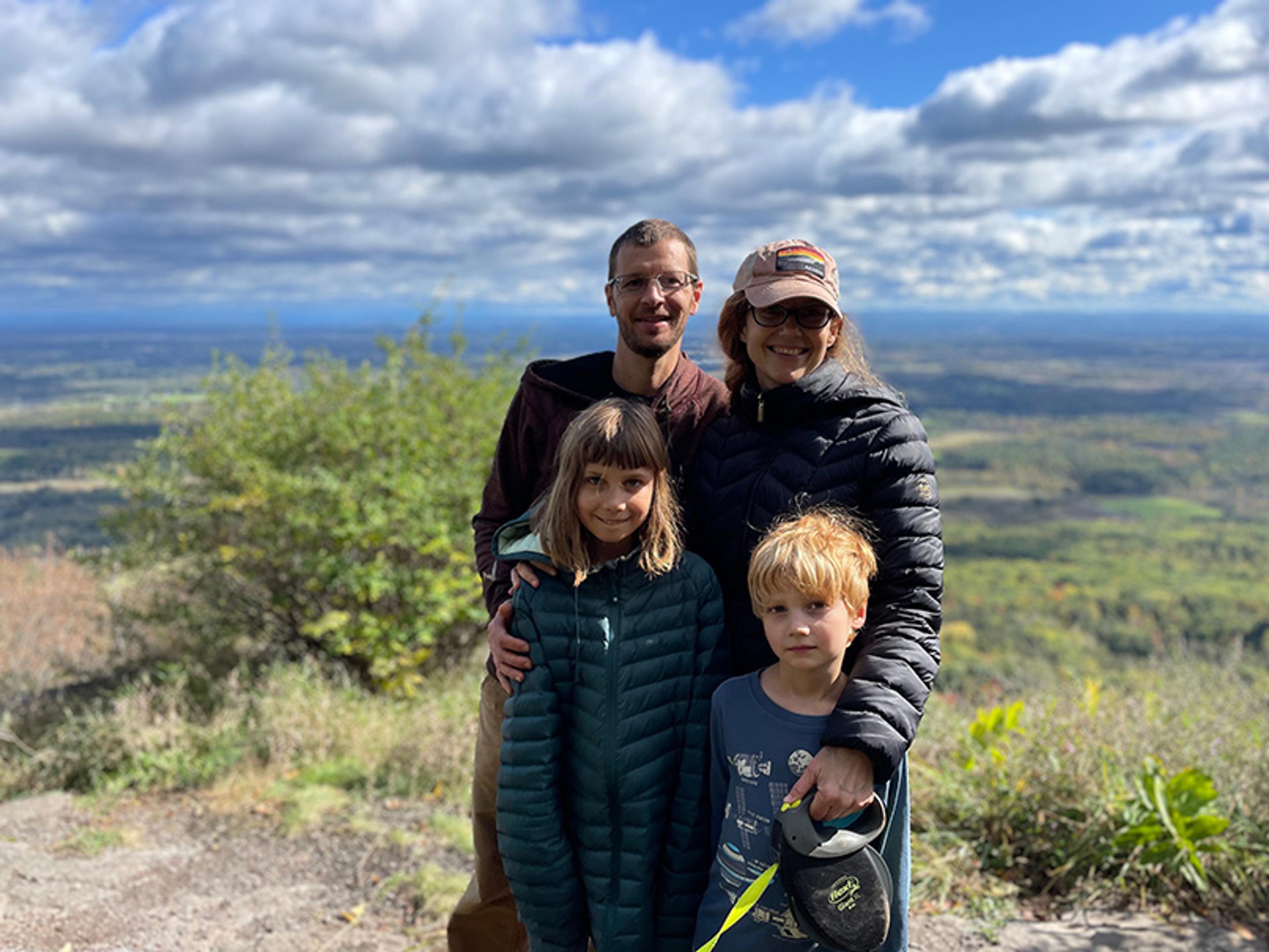 Photo of a family of four, smiling on a hilltop with a scenic landscape and cloudy sky in the background; mother, father and daughter are wearing jackets and mother is wearing a hat.