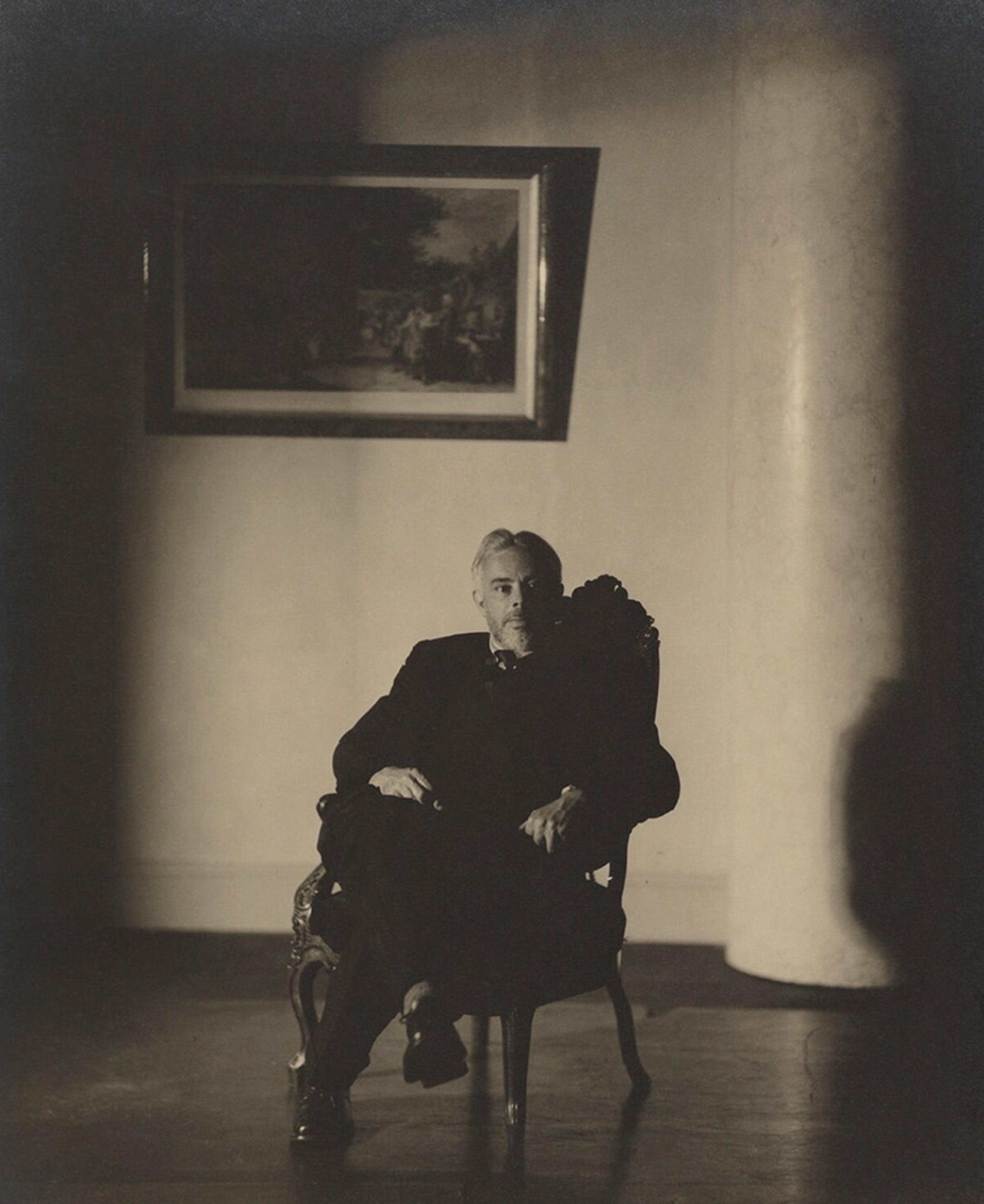 Black and white photo of a man in a suit seated in a dimly lit room with a framed painting and a large pillar behind him.