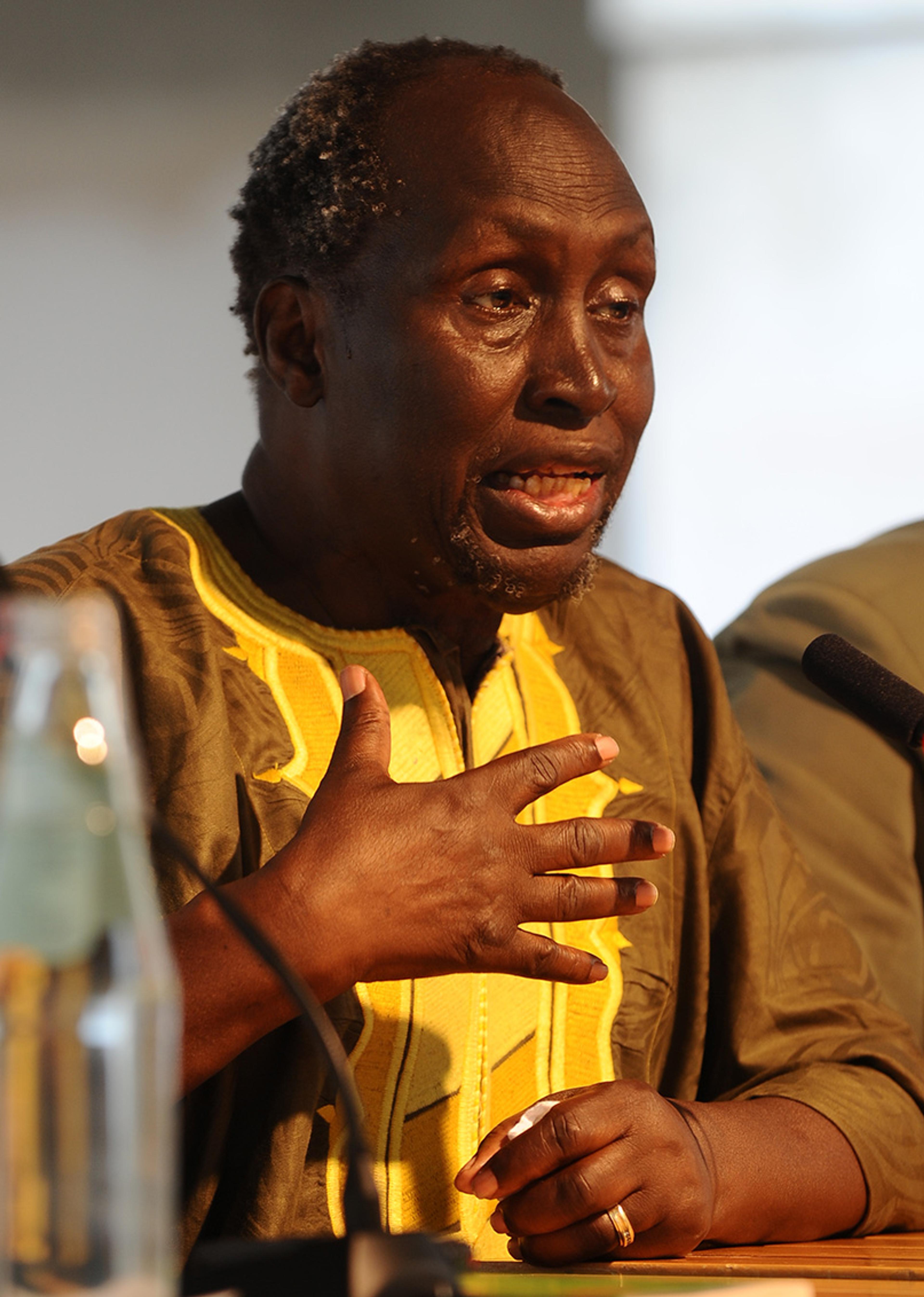 Photo of a man speaking at a podium wearing a yellow and brown traditional outfit, gesturing with his hand.