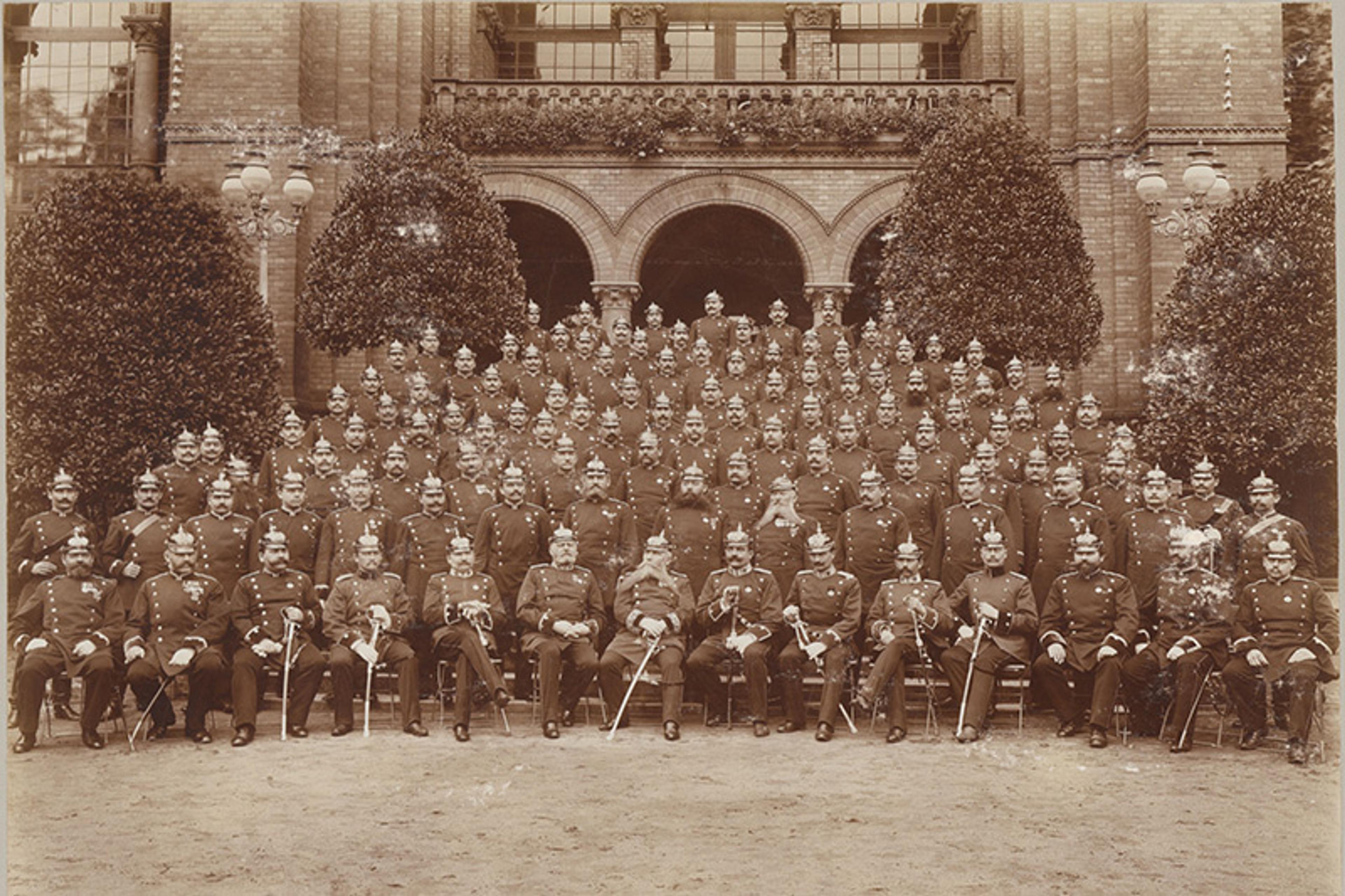 Vintage photo of a large group of uniformed soldiers with spiked pith helmets and swords posed in front of a building with arched entrances and hedges.