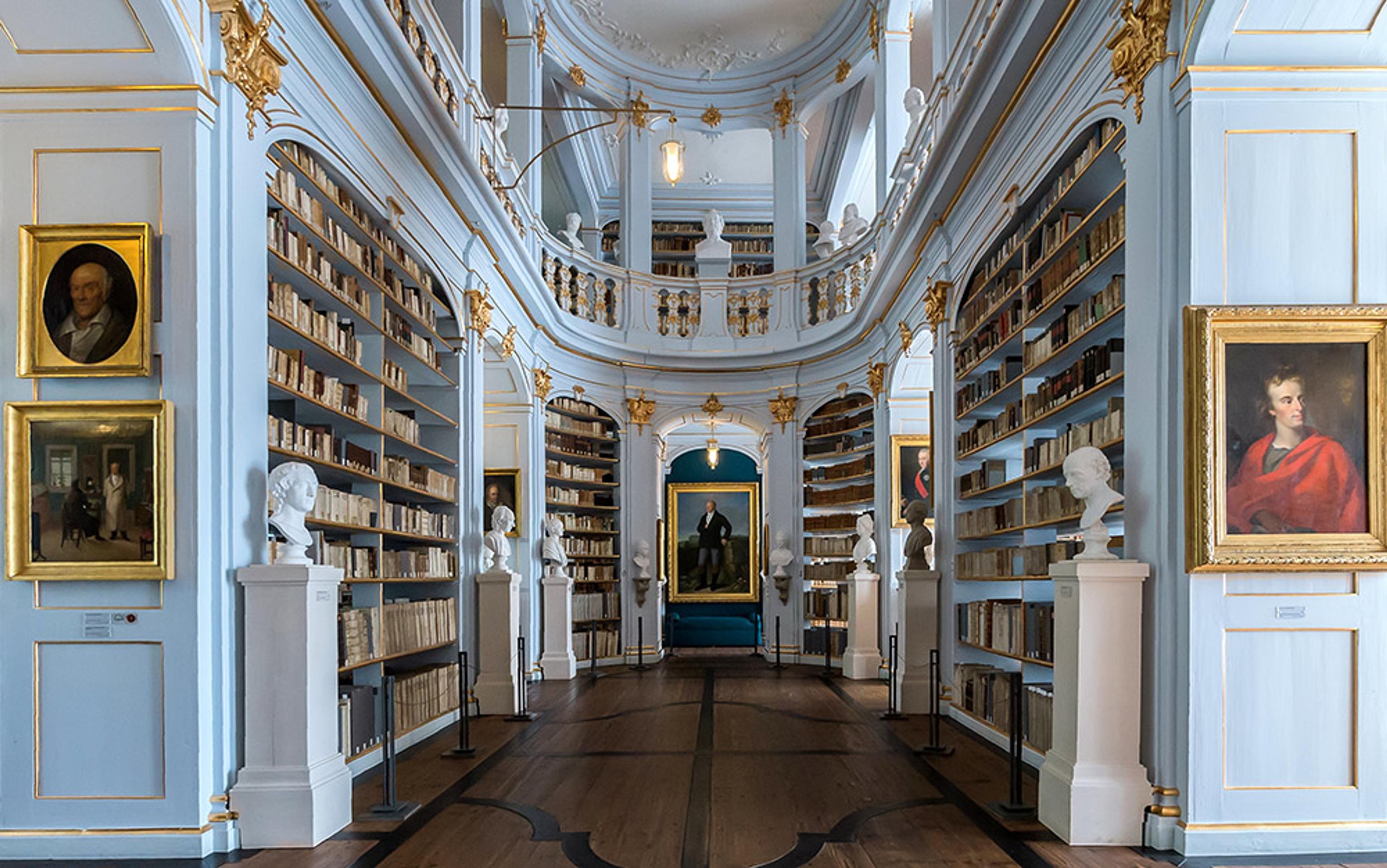 An elegant, historical library with tall bookshelves, framed portraits, busts and a mezzanine level adorned with gold details.