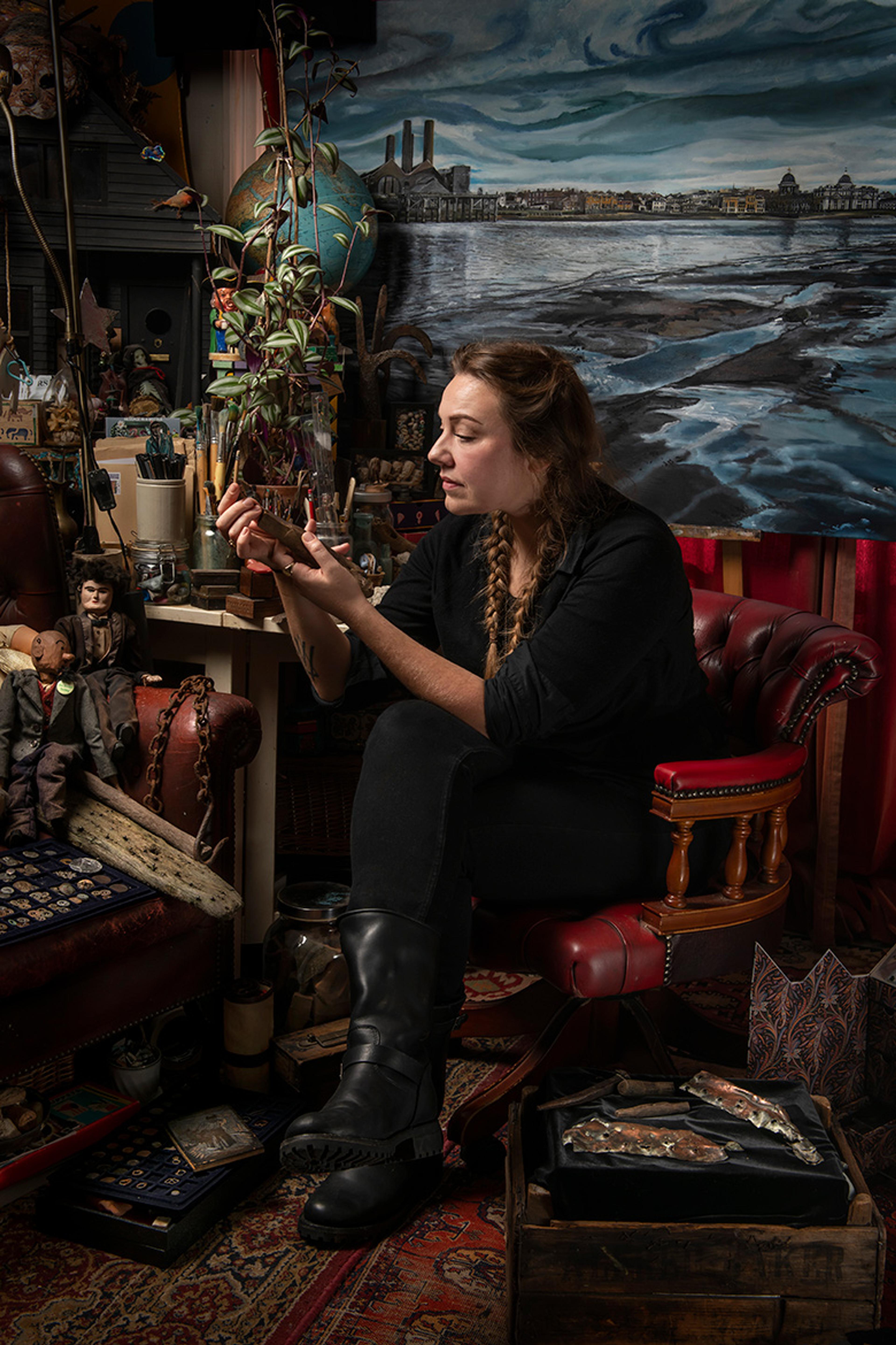 A woman in an art studio surrounded by eclectic objects sitting on a red chair examining a small item.