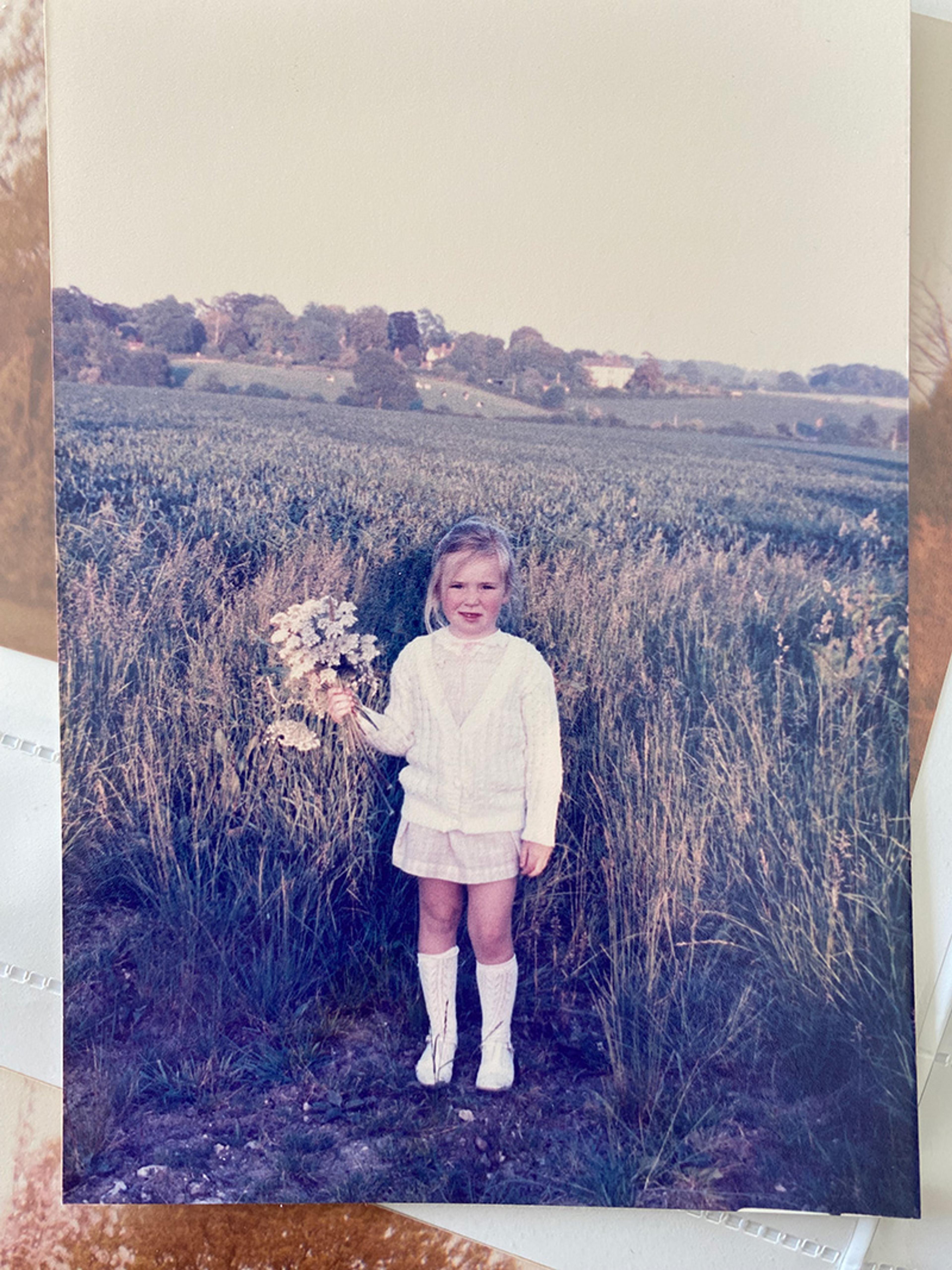 Photo of a young girl in a field holding wildflowers, wearing a white jumper and skirt with trees and hills in the background.