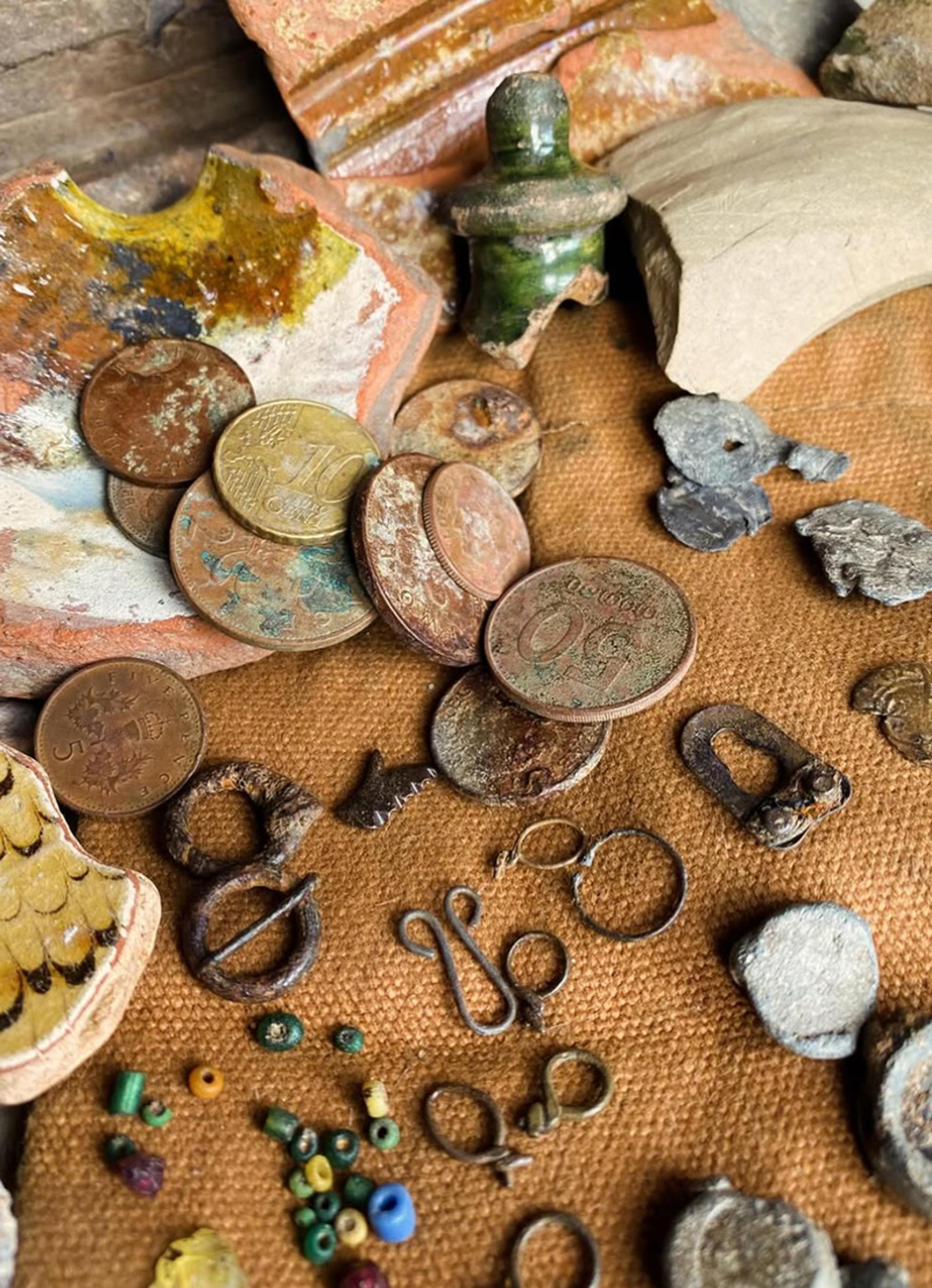 Coins, pottery shards, beads and metal artefacts on a textured surface, showing various aged and weathered items.