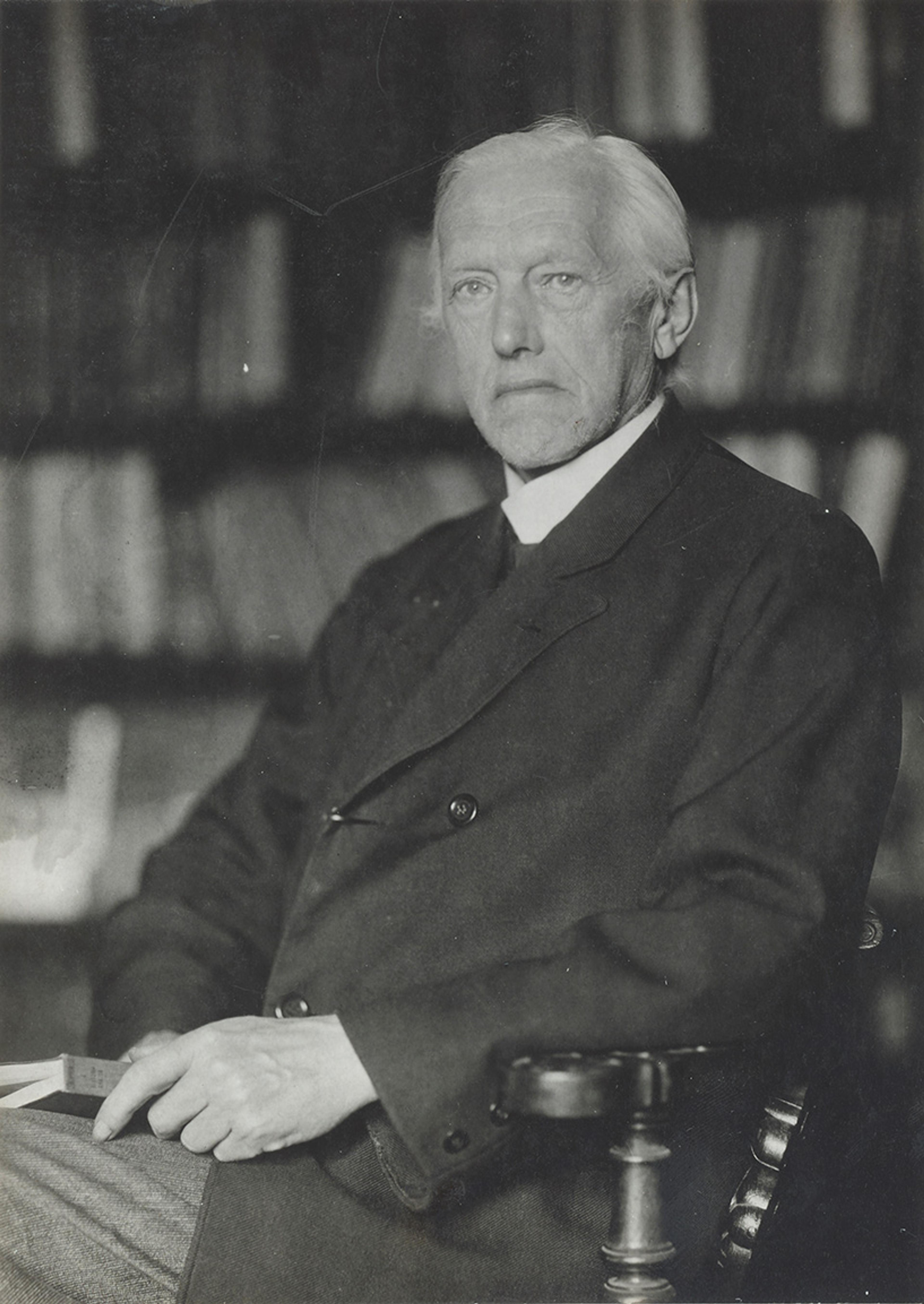 Black and white photo of an elderly man in a suit sitting on a chair in front of bookshelves.