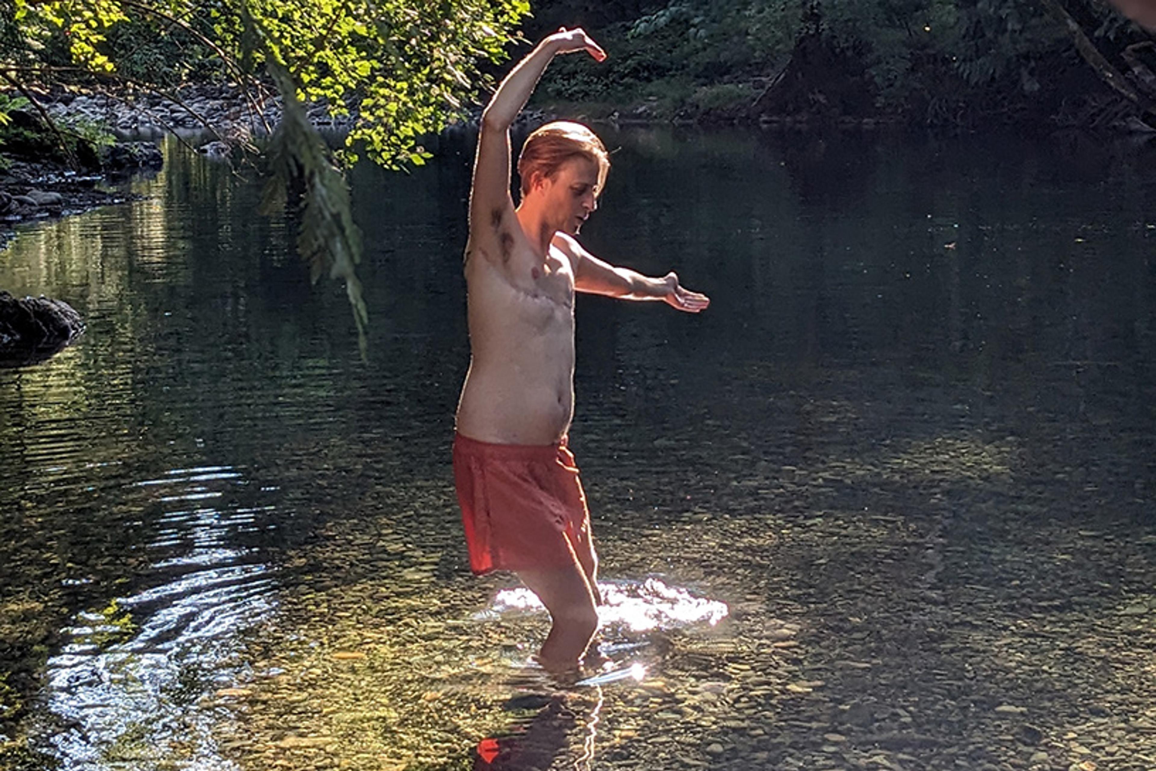 A person in red shorts with arms raised, standing in a sunlit shallow river surrounded by trees.