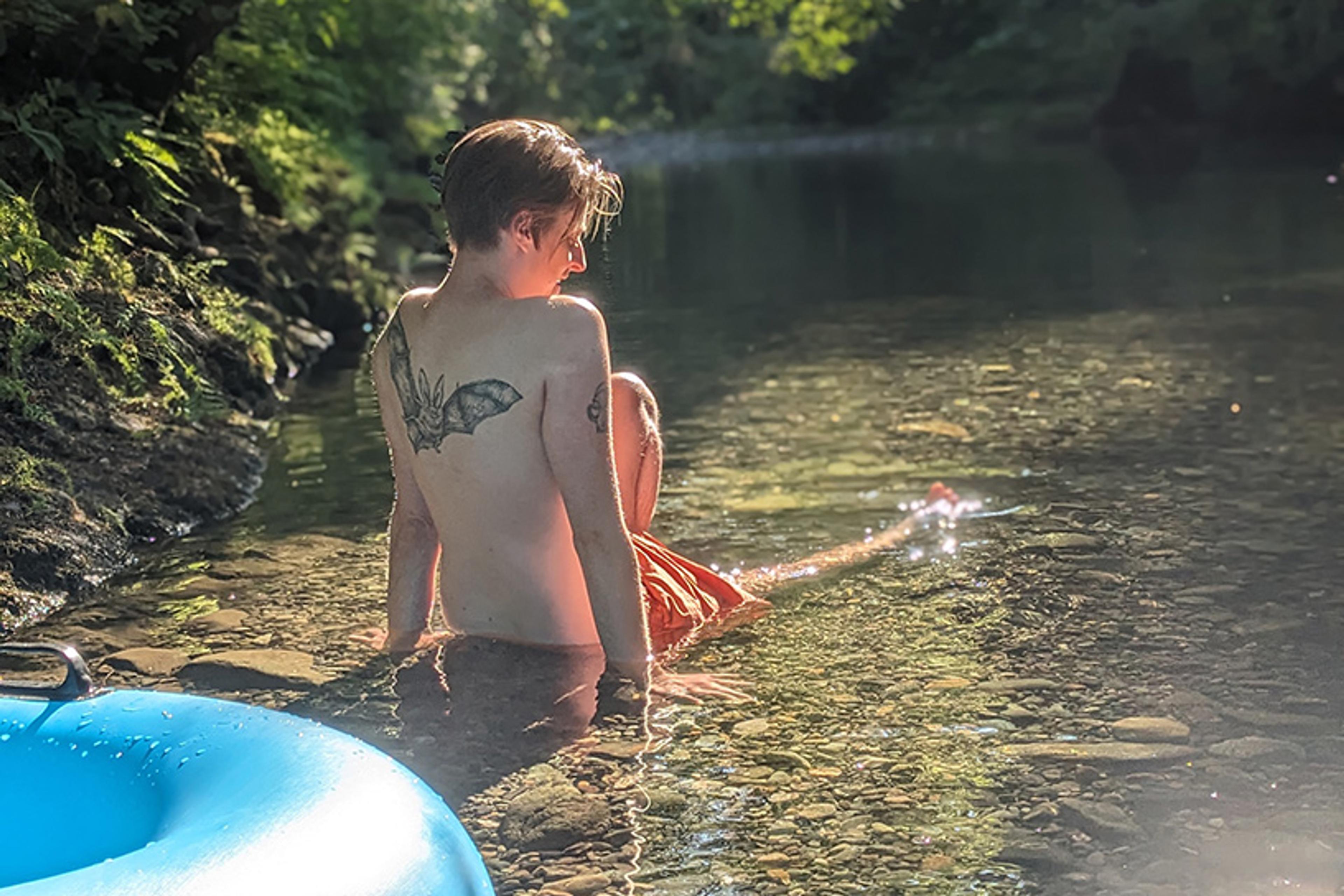 A person with tattoos sitting in a sunlit river, near a blue inflatable ring, surrounded by trees.
