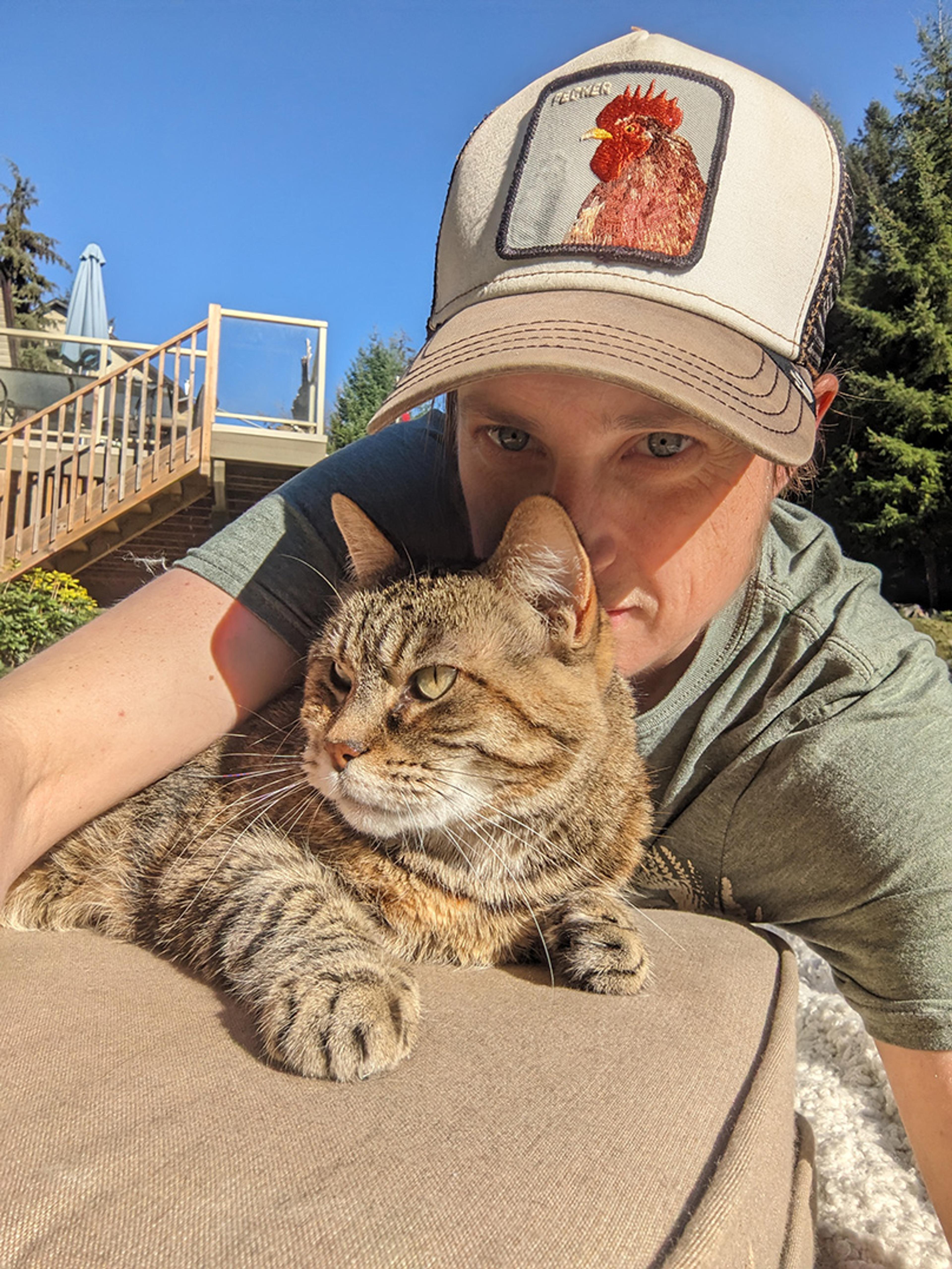 A person wearing a cap with a rooster logo, cuddling a tabby cat outdoors in sunny weather.