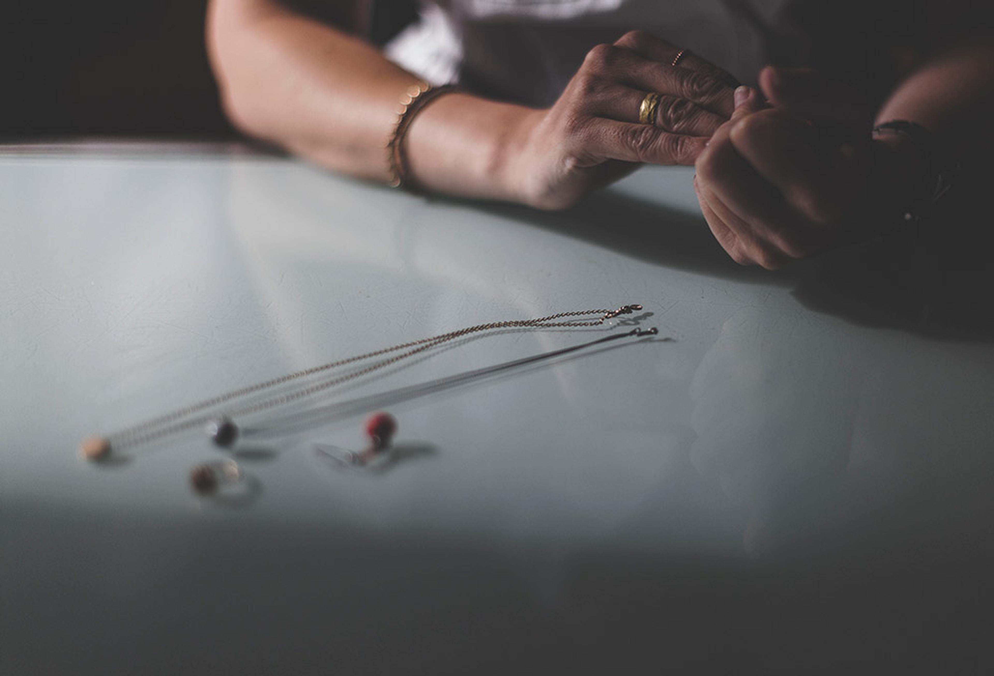 A person’s hands on a table with jewellery, including a necklace and rings, lying on the tabletop.