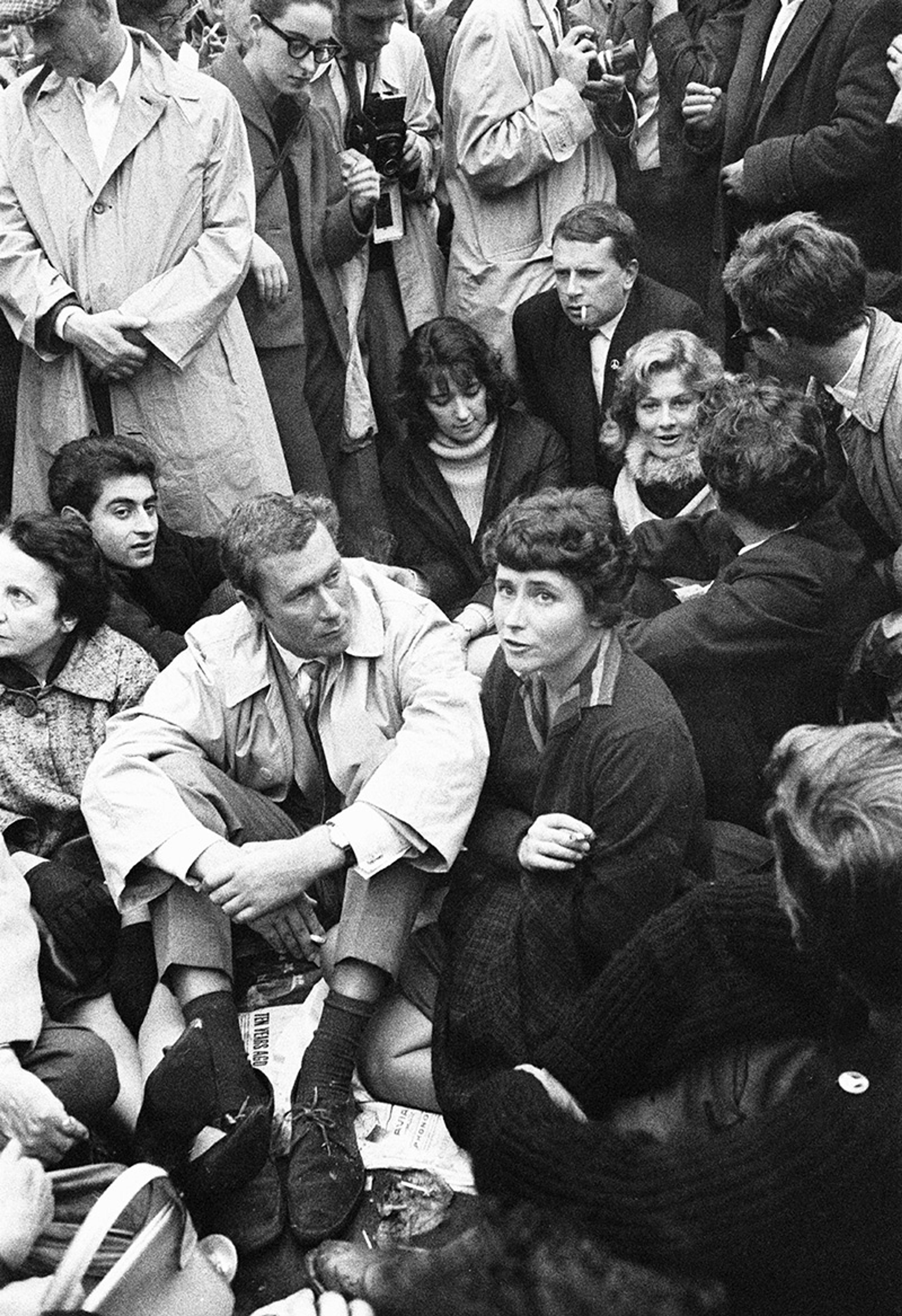 Black and white photo of a group of people sitting and standing close together, some wearing coats, one person smokes a cigarette.