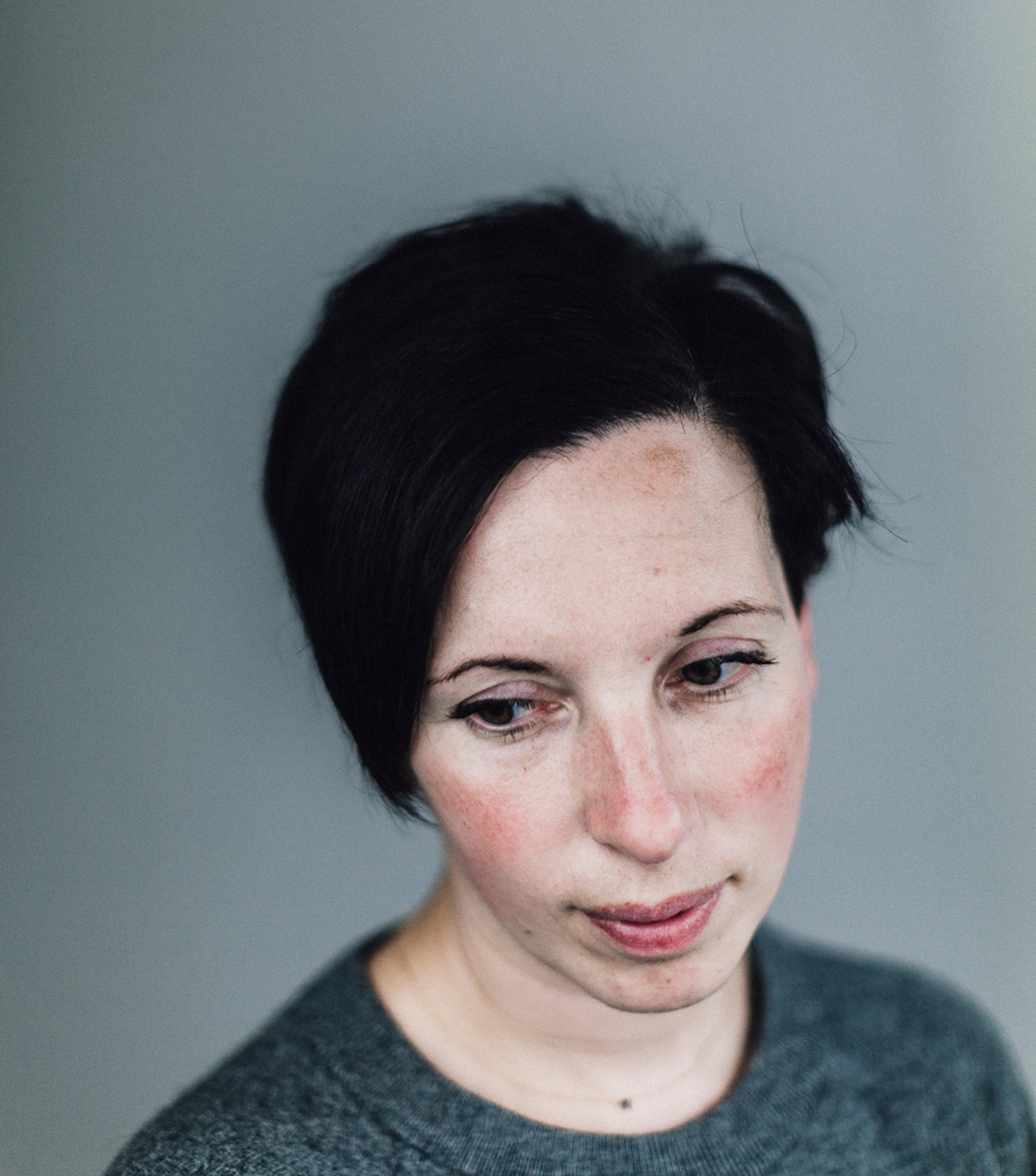A woman with a short dark hairstyle, wearing a grey top, looking down with a neutral expression. Grey background.