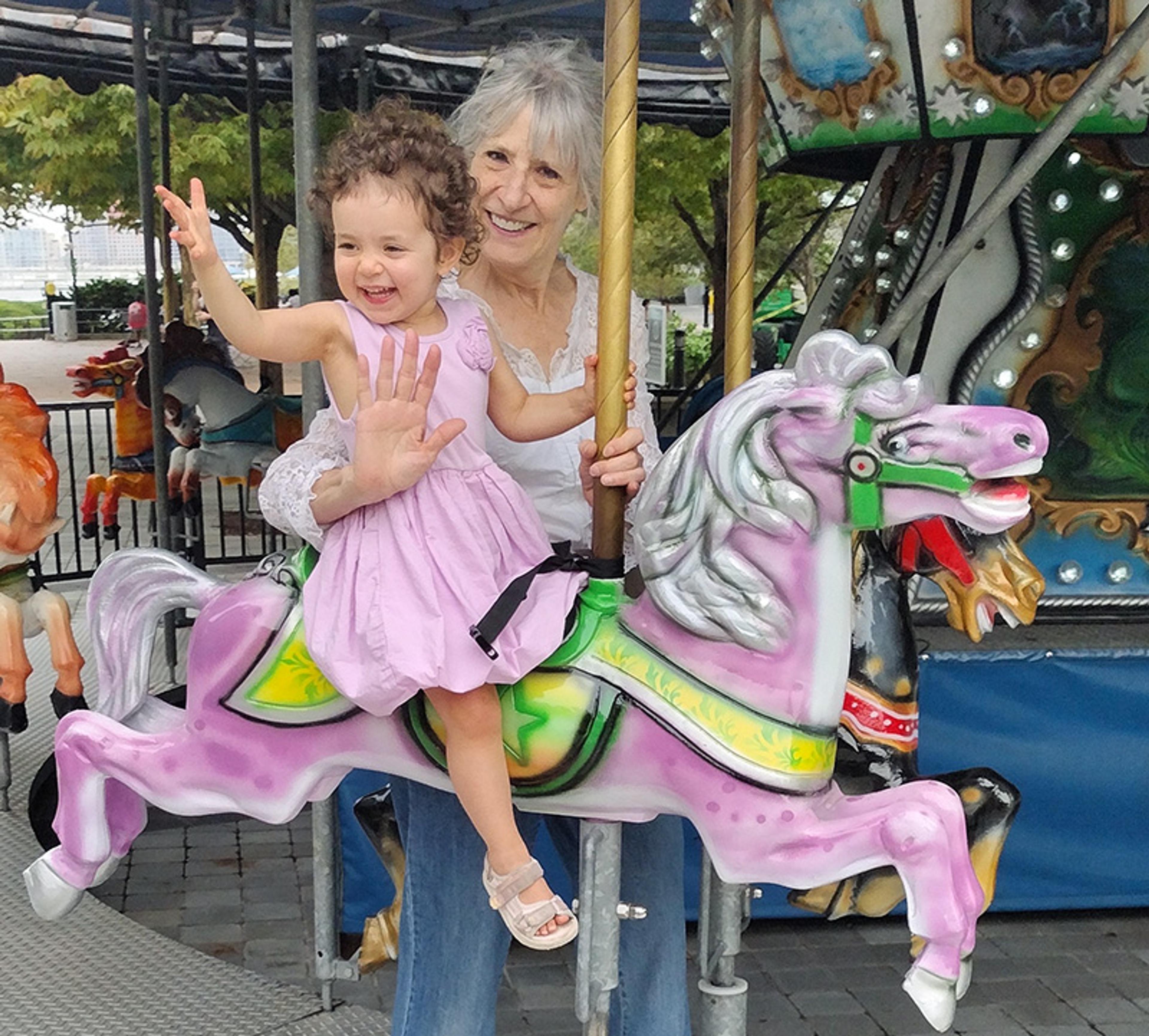 A smiling woman and child on a carousel horse at an amusement park, with trees in the background.
