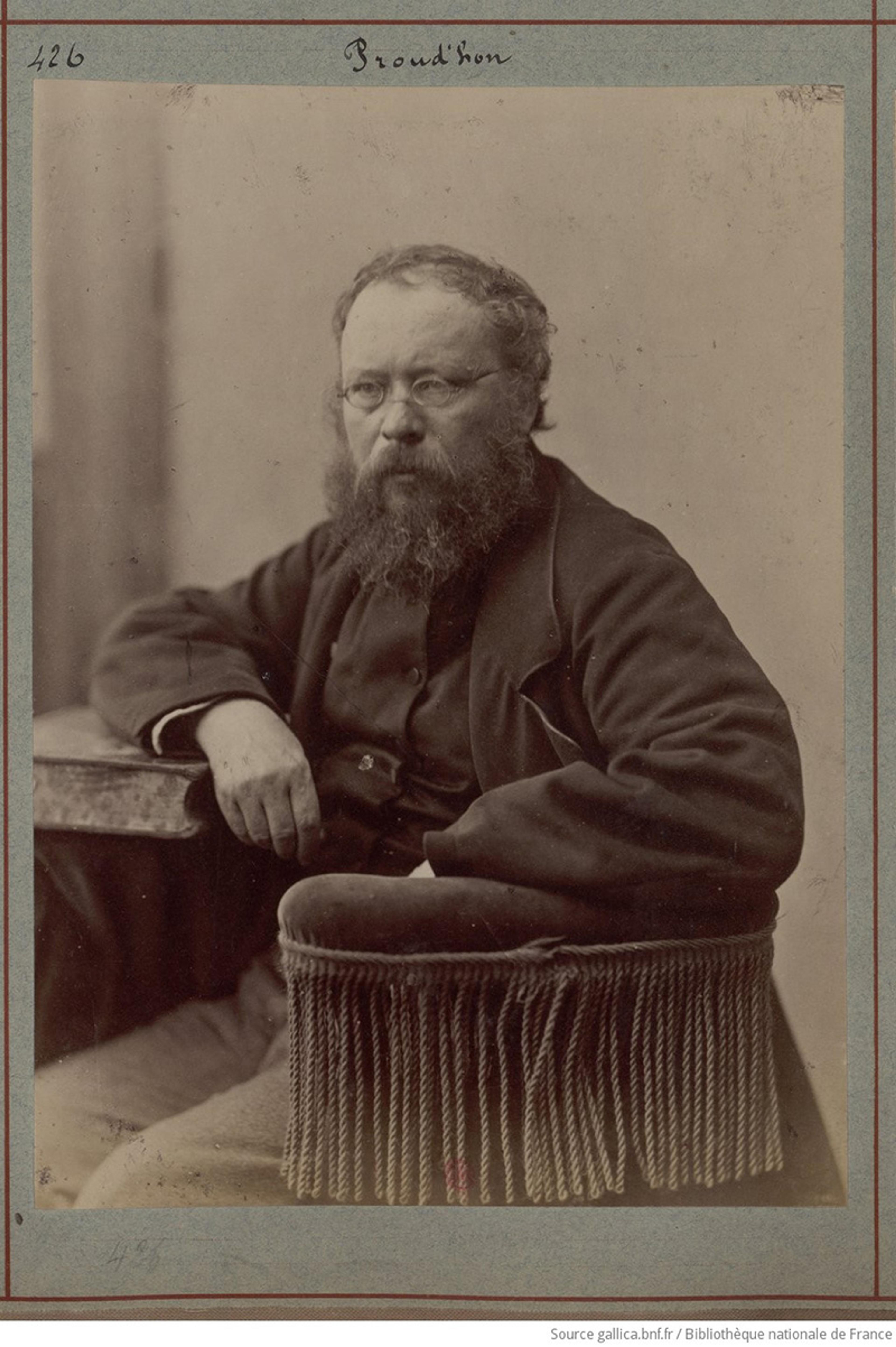 Sepia-toned photo of a seated man with a beard and glasses, resting on a chair with fringe, looking thoughtful.
