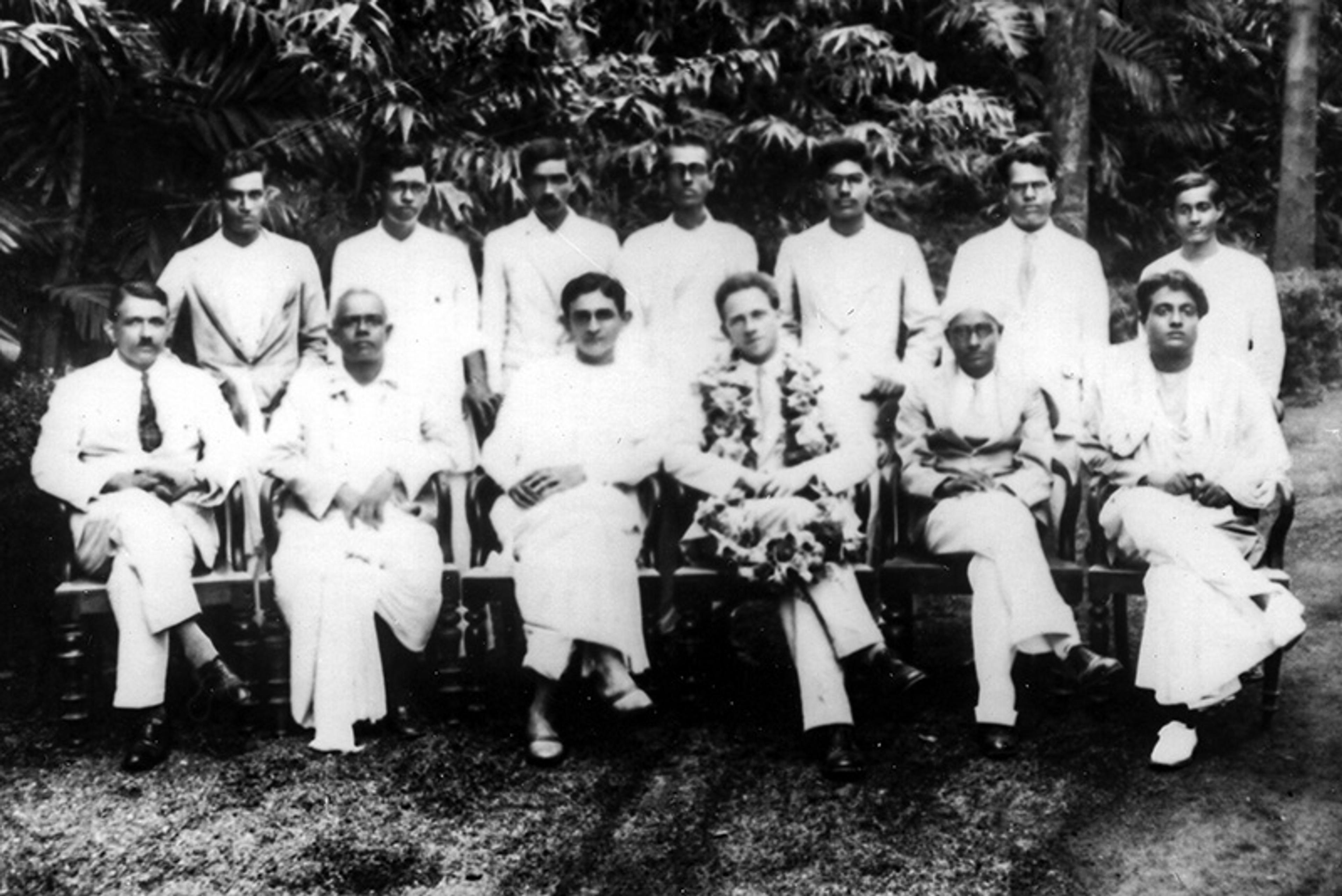 Black and white photo of a group of men in traditional and Western attire, seated and standing together outdoors.