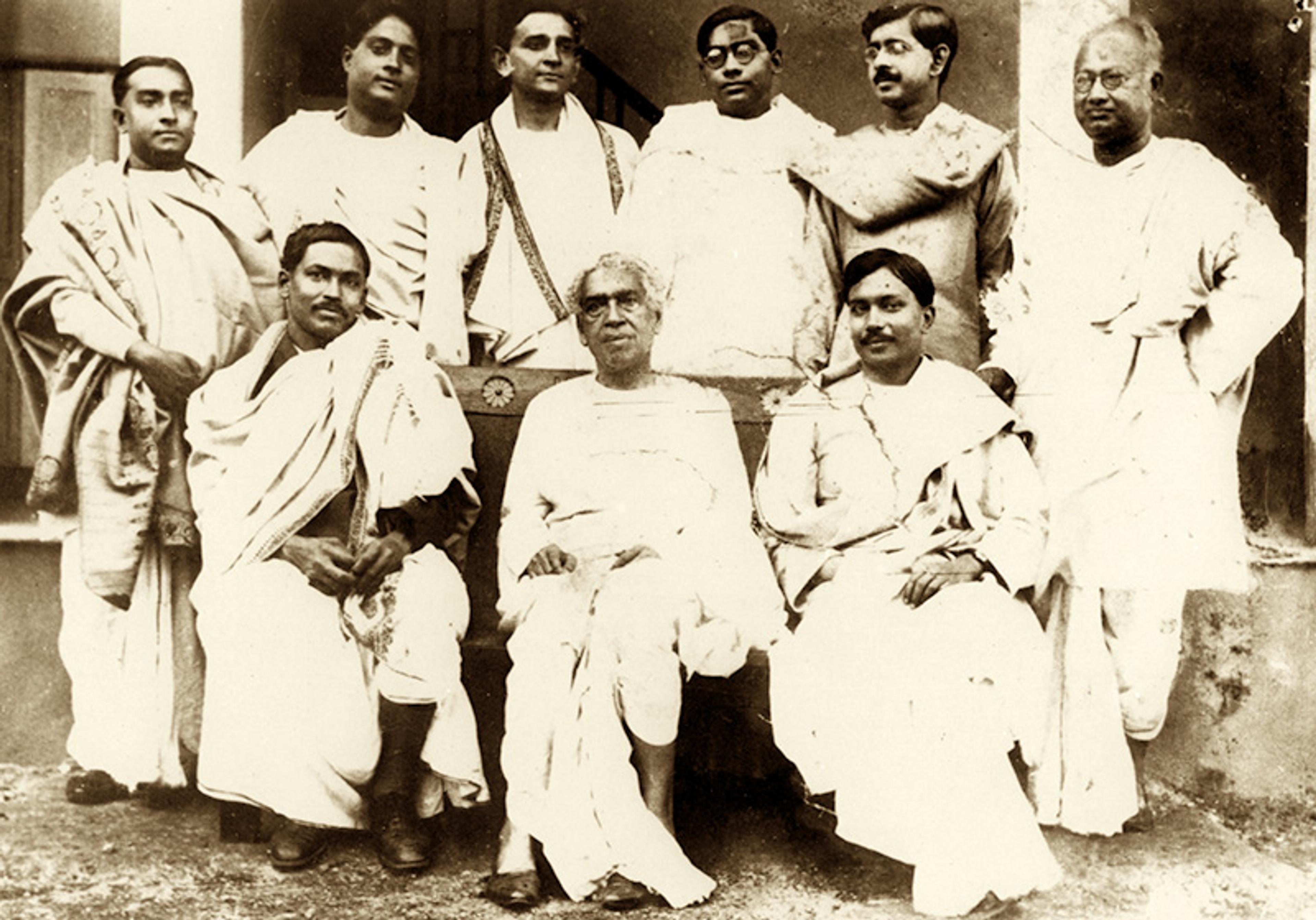 Vintage photo of a group of nine men in traditional Indian attire, with three seated in front and six standing behind.