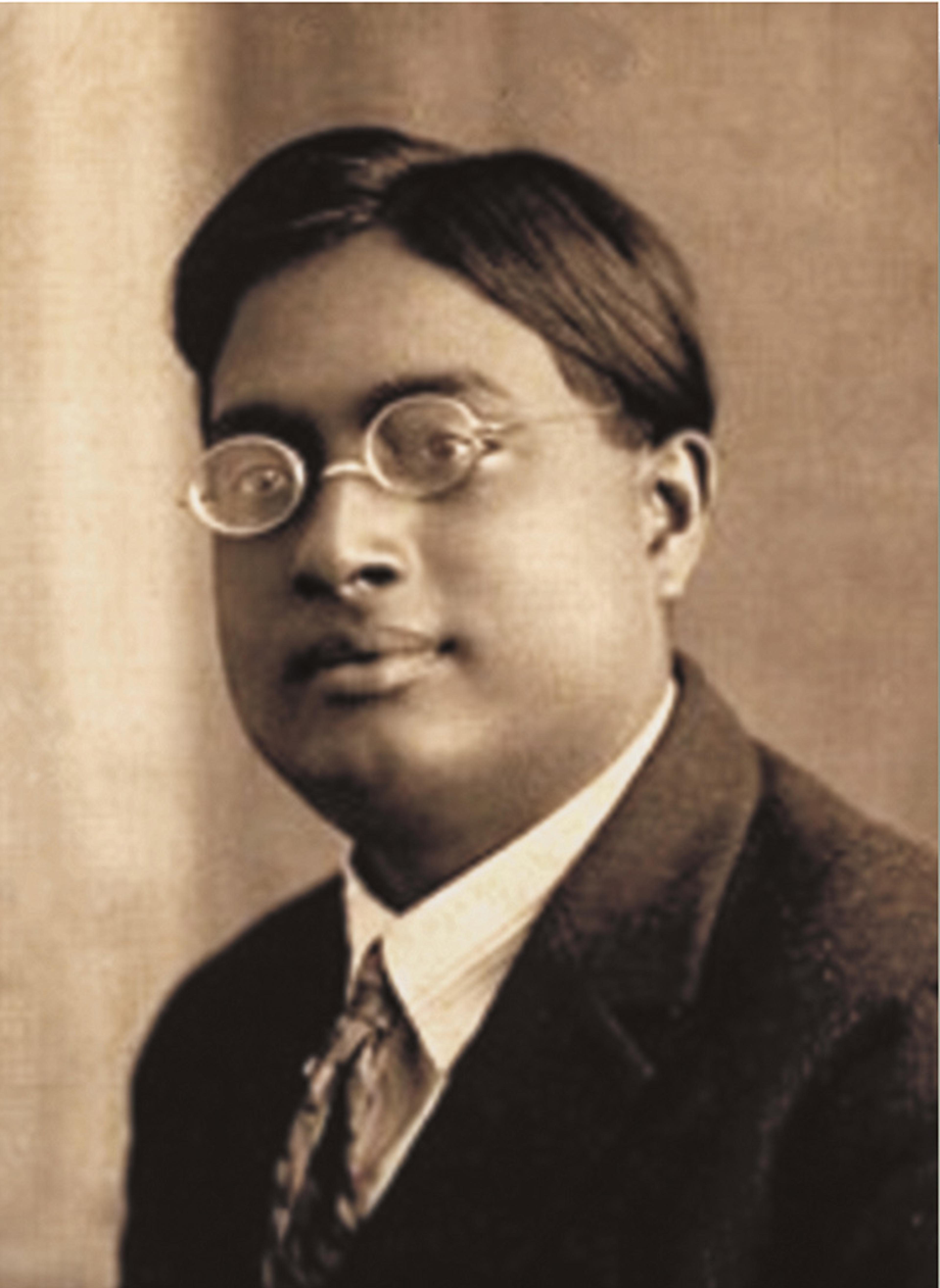 Sepia-toned photo of a bespectacled man in a suit with a tie, head slightly tilted, early 20th century style.