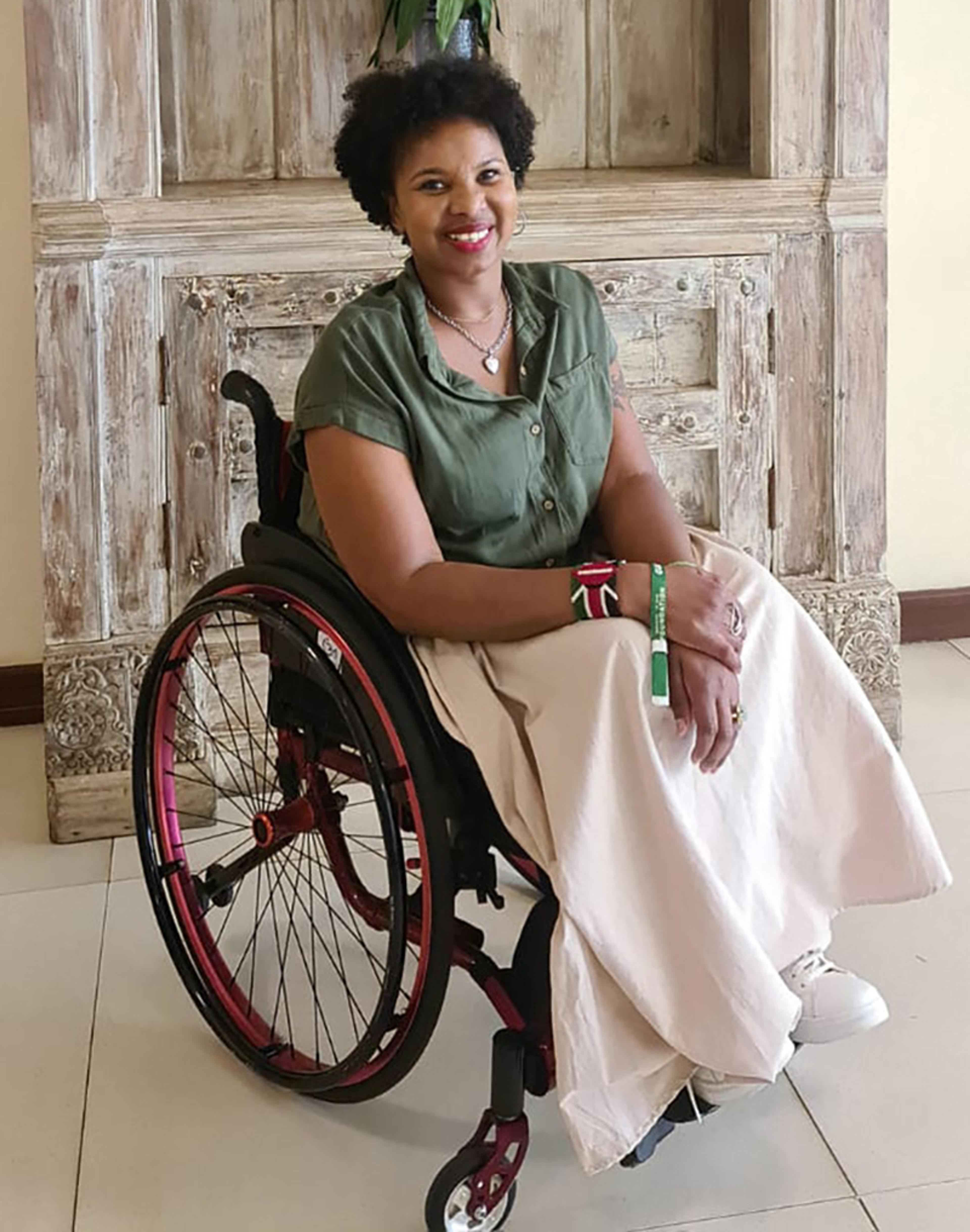 Photo of a smiling person in a wheelchair wearing a green shirt and beige skirt against a rustic wooden backdrop.