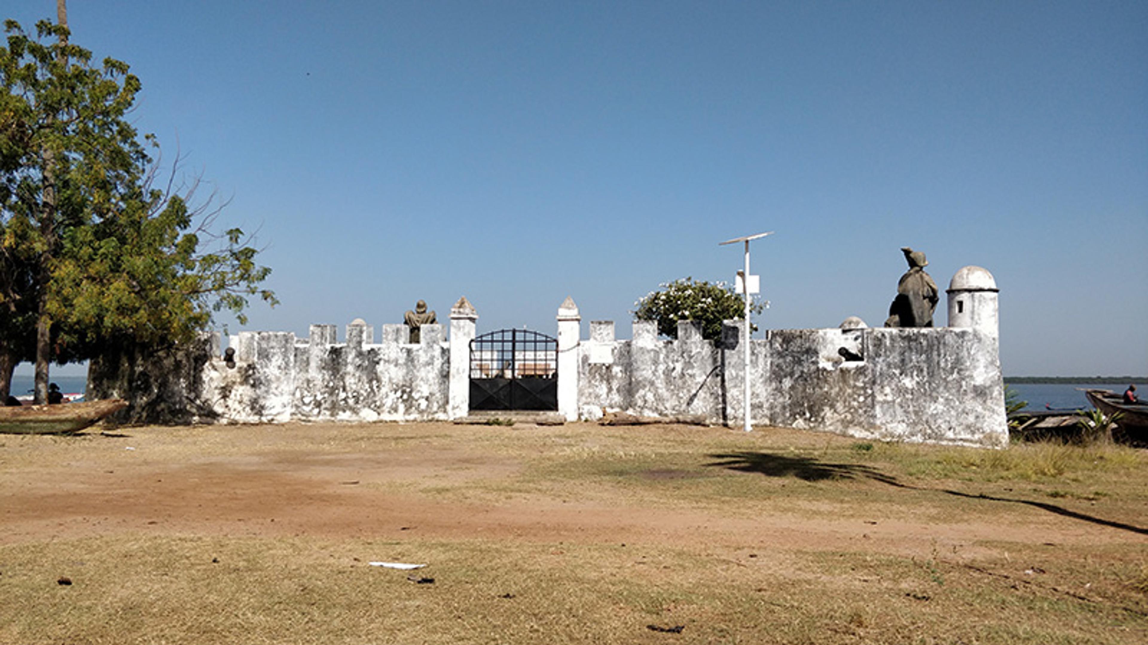 Photo of an old fort with weathered white walls, a gate, and statues under a clear blue sky with surrounding trees and grass.