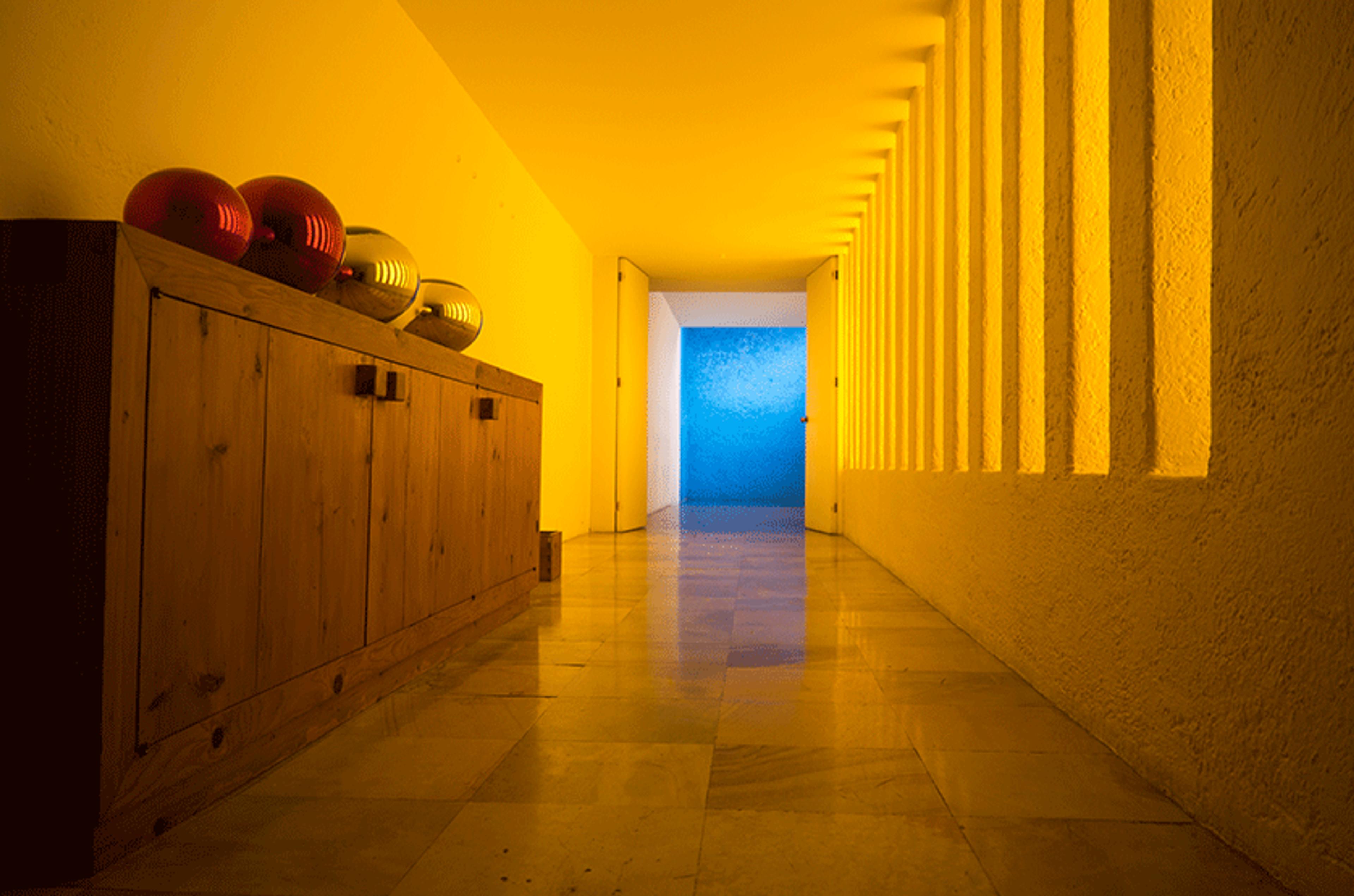 Photo of a hallway with yellow lighting, wooden cabinet with vases on left, blue wall at end. Floor is reflective marble.
