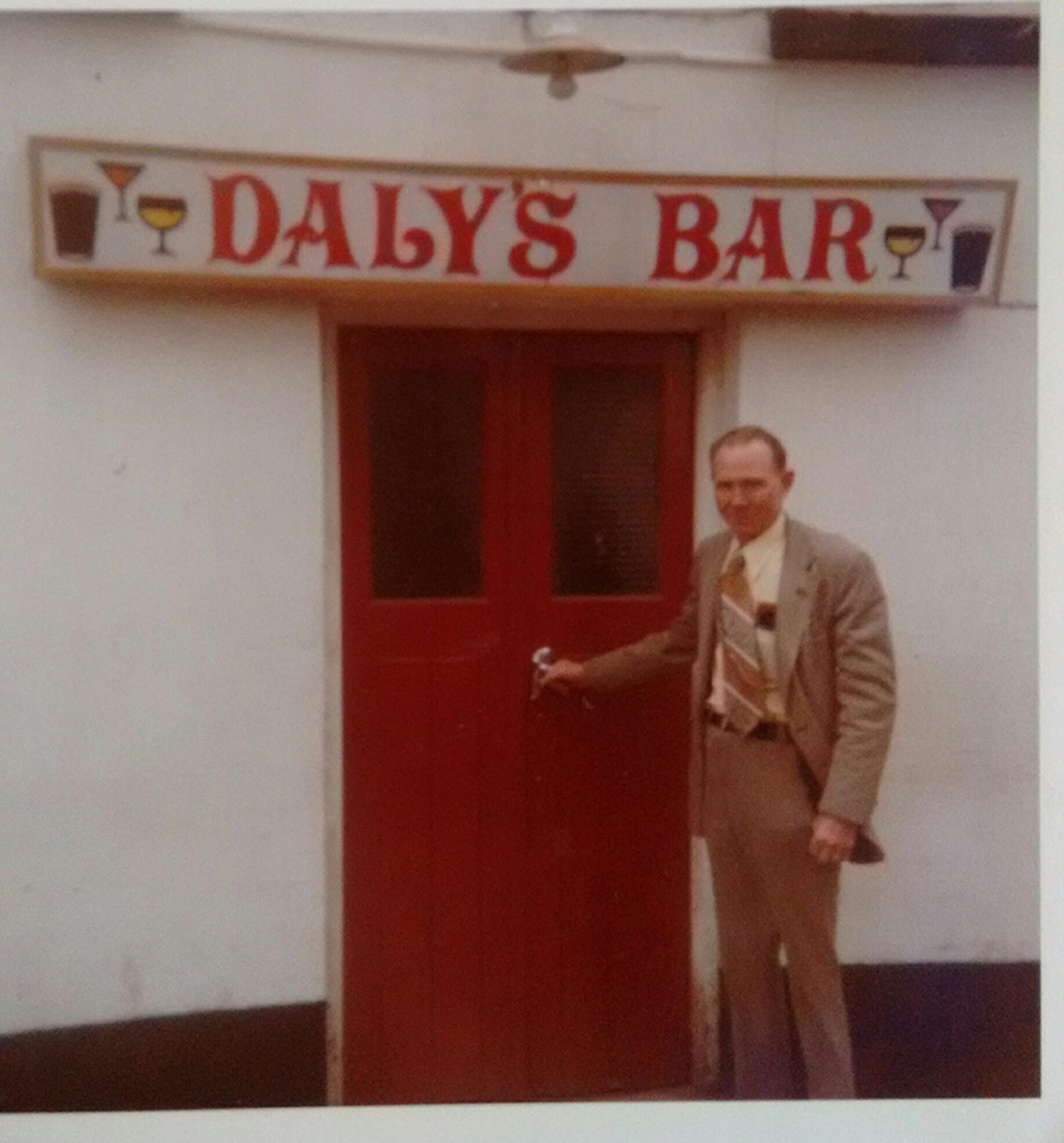 A man in a suit standing by the red door of Daly’s Bar, with a sign above depicting drinks.