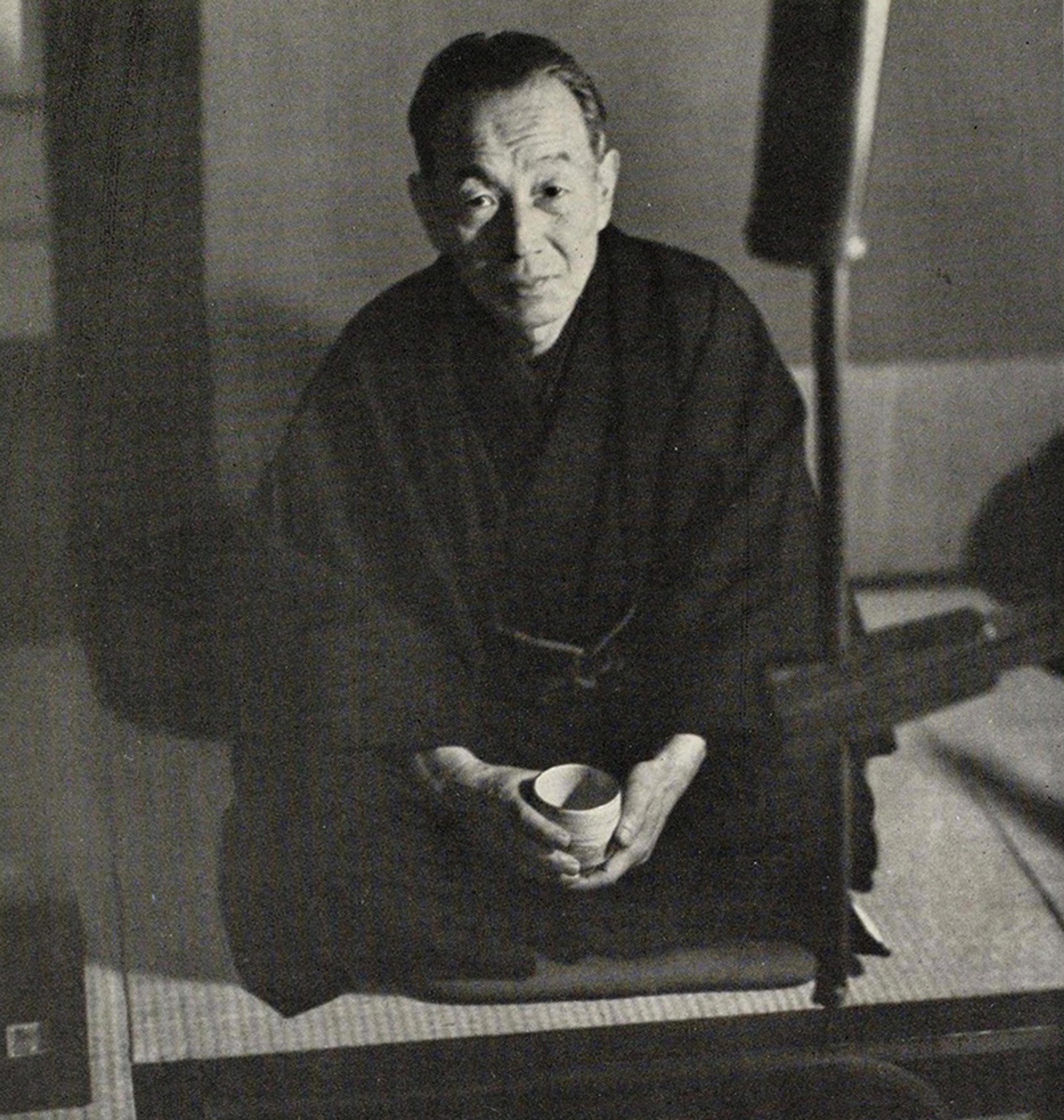 Black and white photo of a seated person in traditional attire holding a cup indoors.