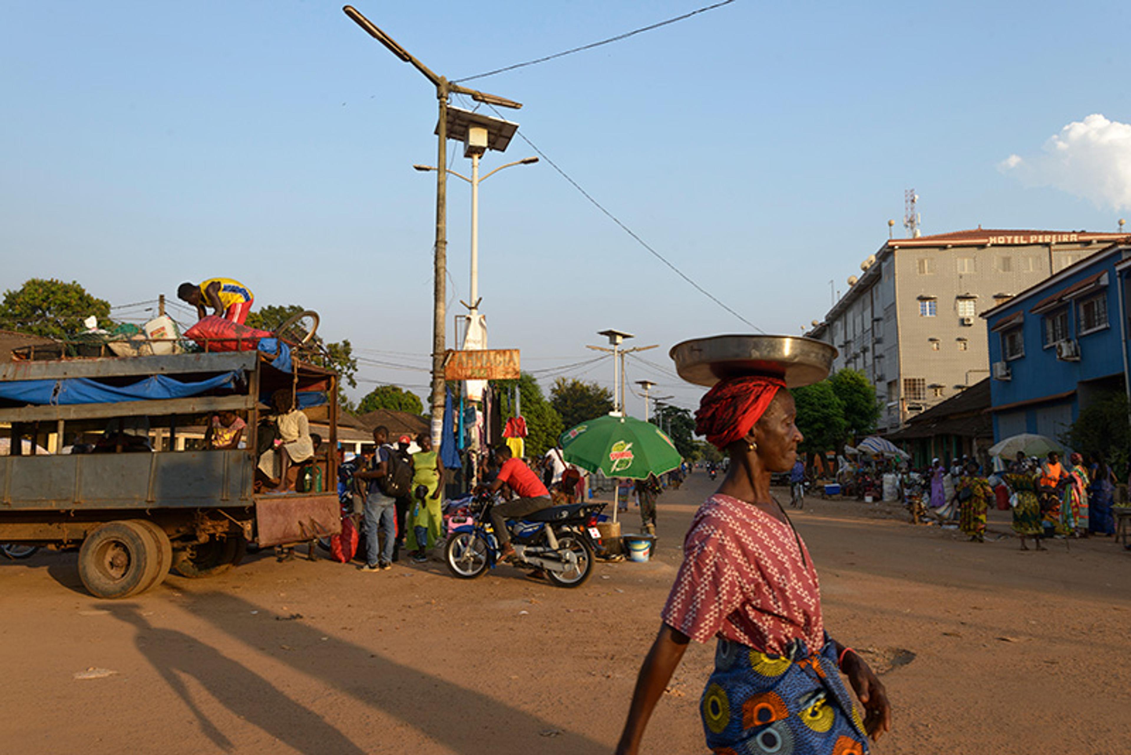 Photo of a busy street scene in sunlight with people, a woman carrying a basin on her head, motorcycles and a truck.